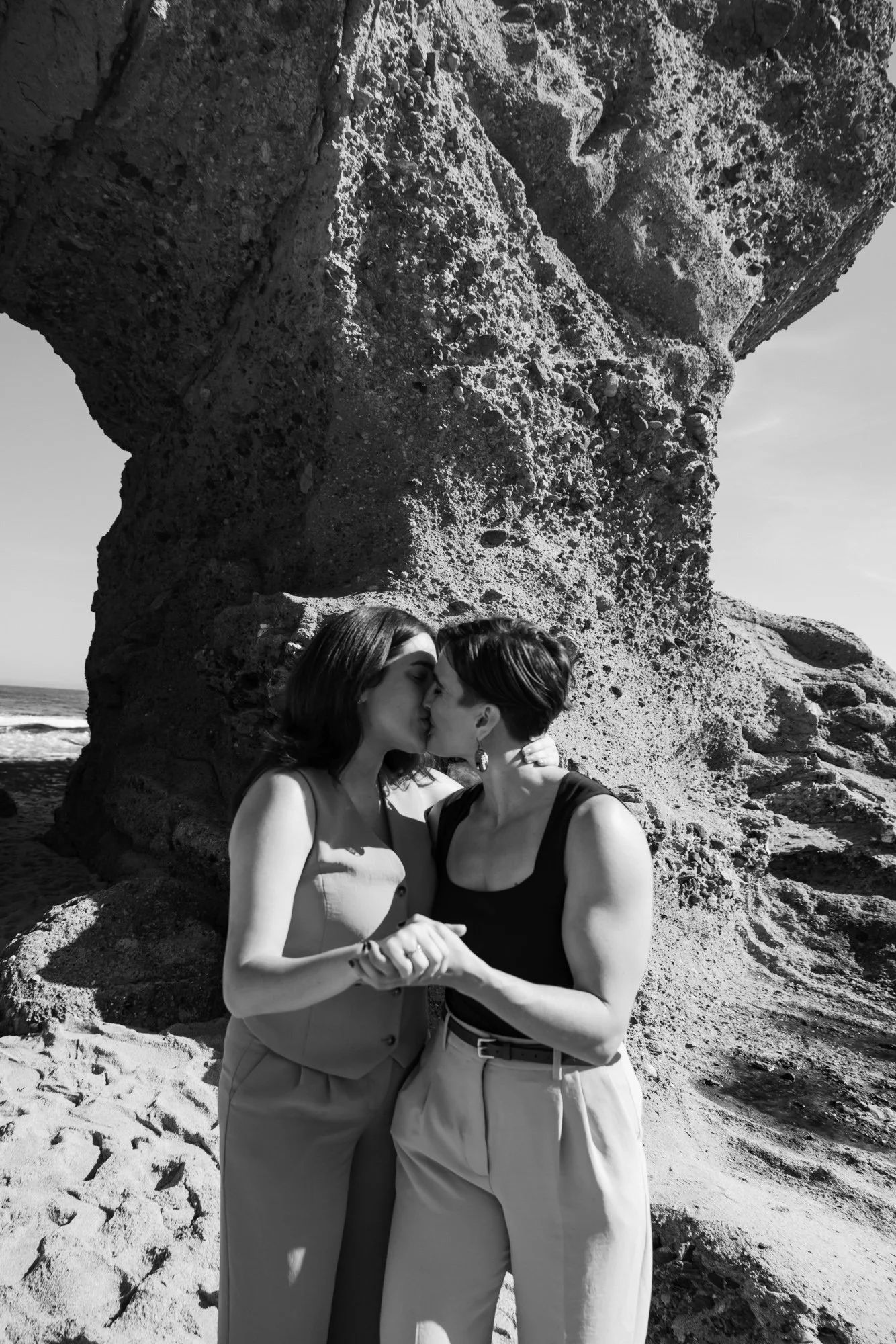 Two women sharing a kiss on a beach with large rock formations in the background, photograph in black and white.