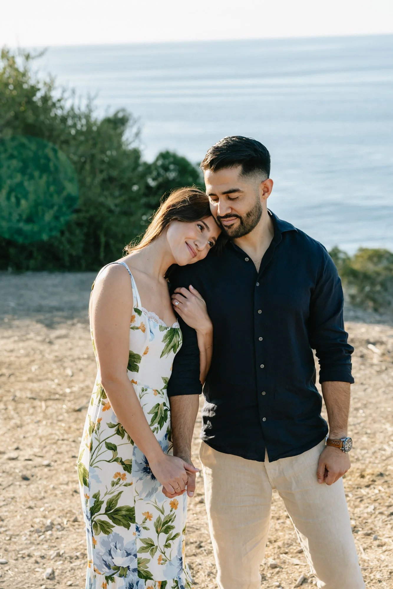 A couple holding hands and embracing by the water, with the woman resting her head on the man's shoulder, on a sunny day at the beach.