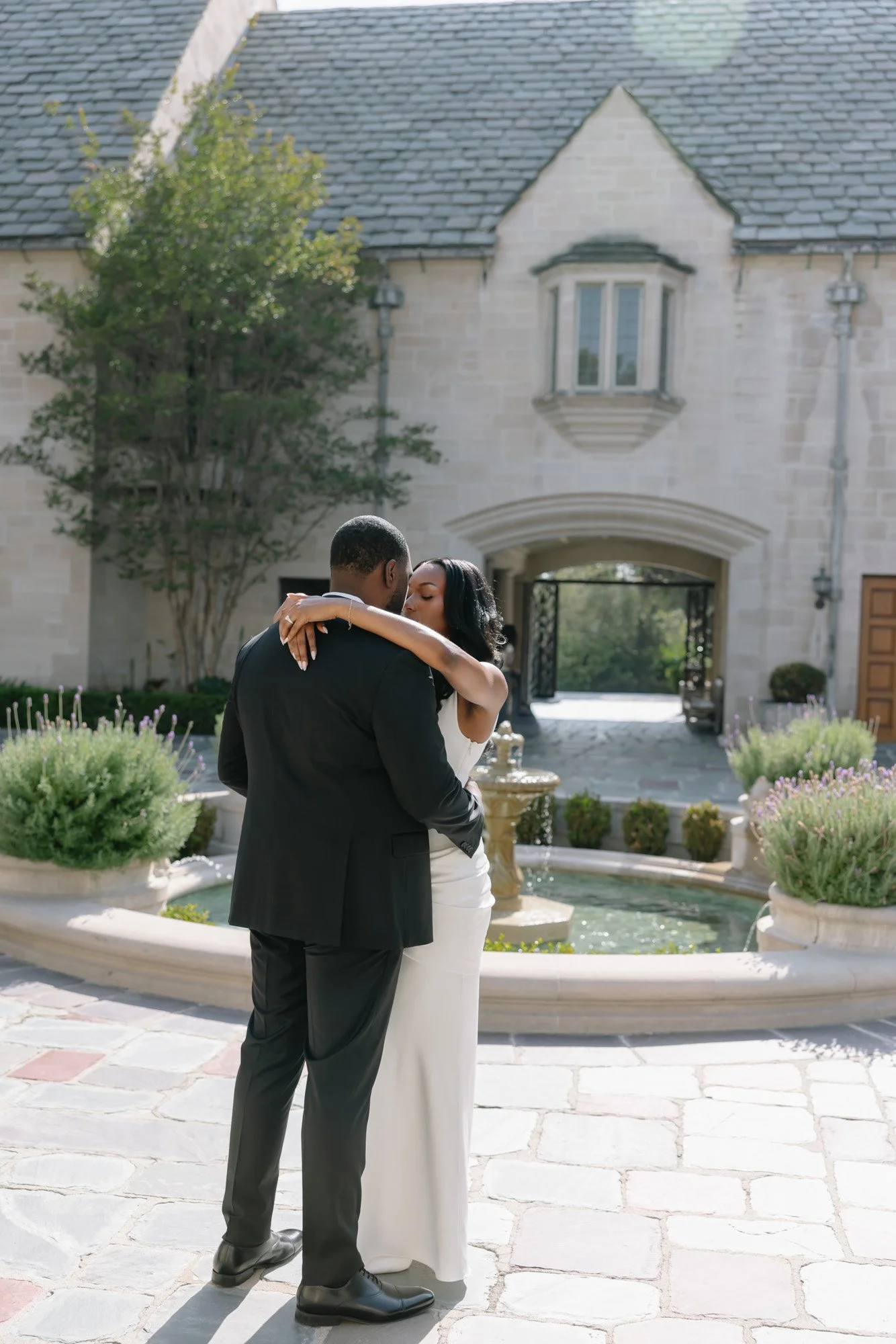 A couple dressed in wedding attire kissing outdoors in front of a stone building with a fountain and potted plants.
