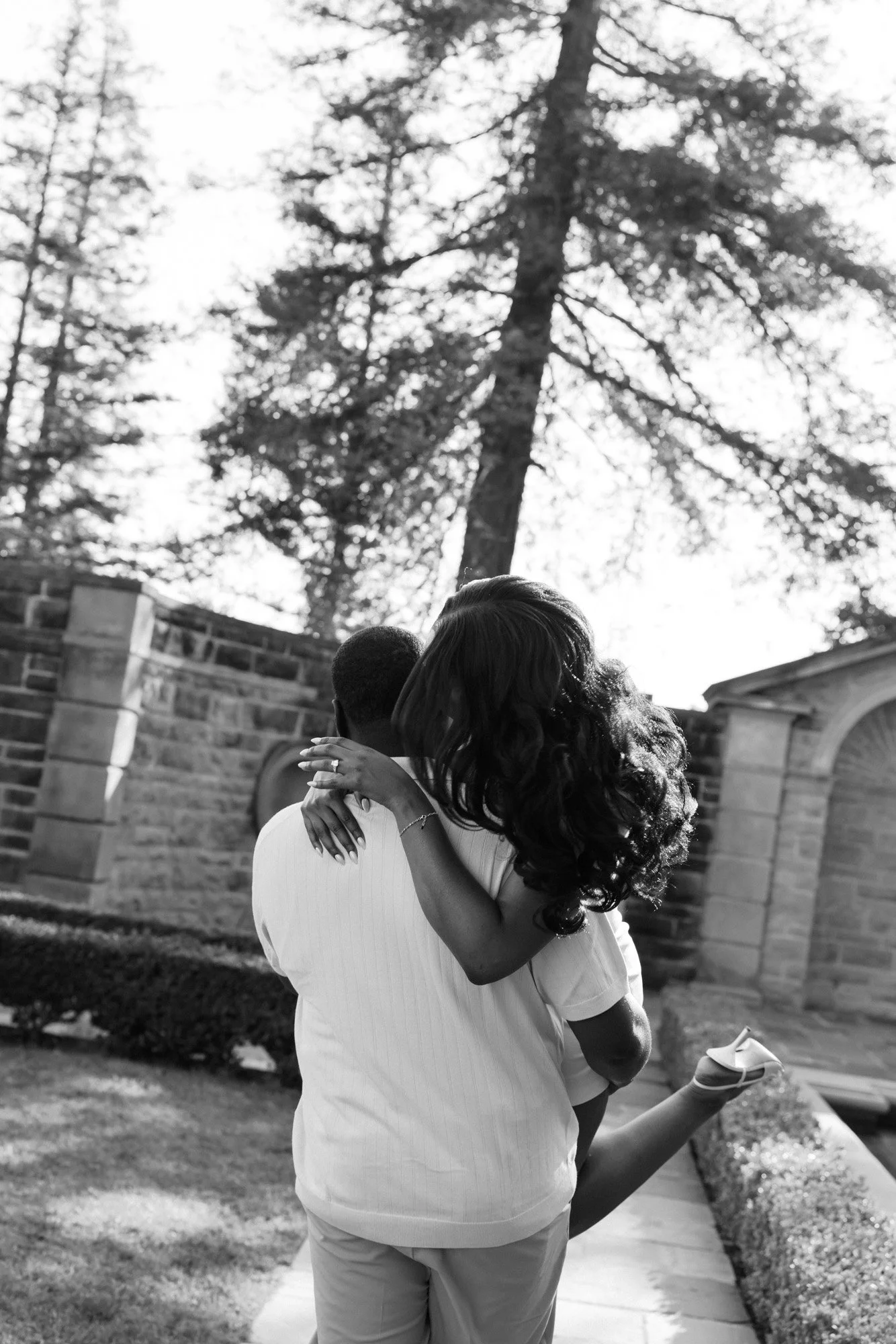 A couple embraces outdoors on a sunny day, the woman with long, curly hair and the man with short hair, in front of a brick wall and a large tree.
