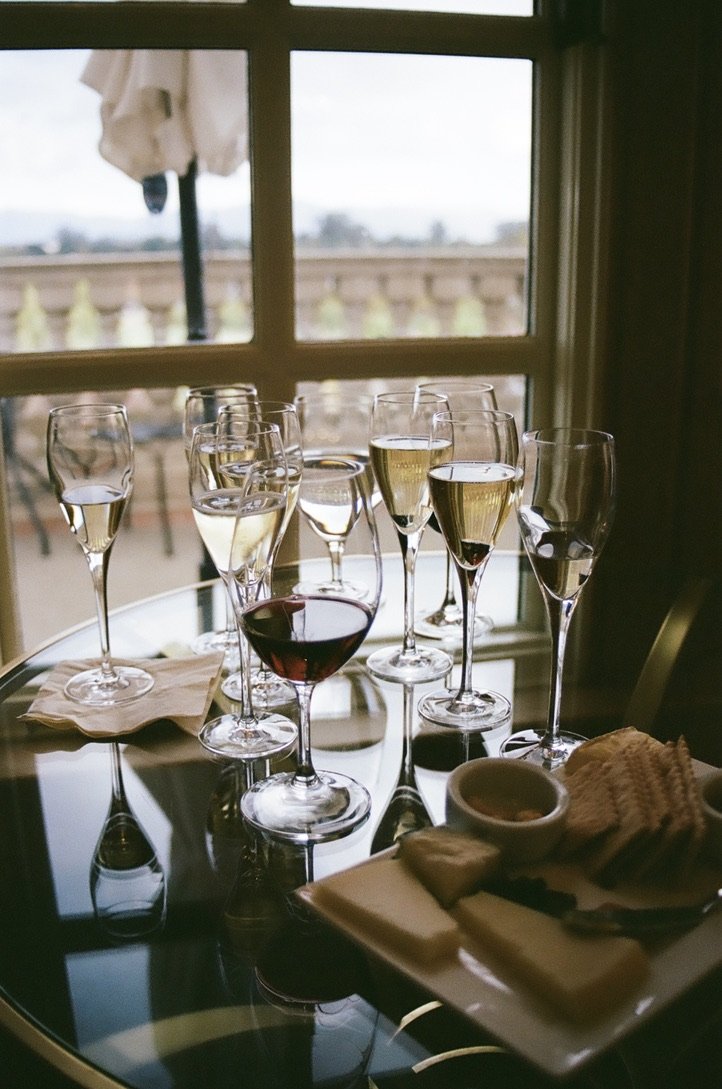 Several glasses of assorted wine on a glass table near a window, with a plate of snacks and a bowl of dip.