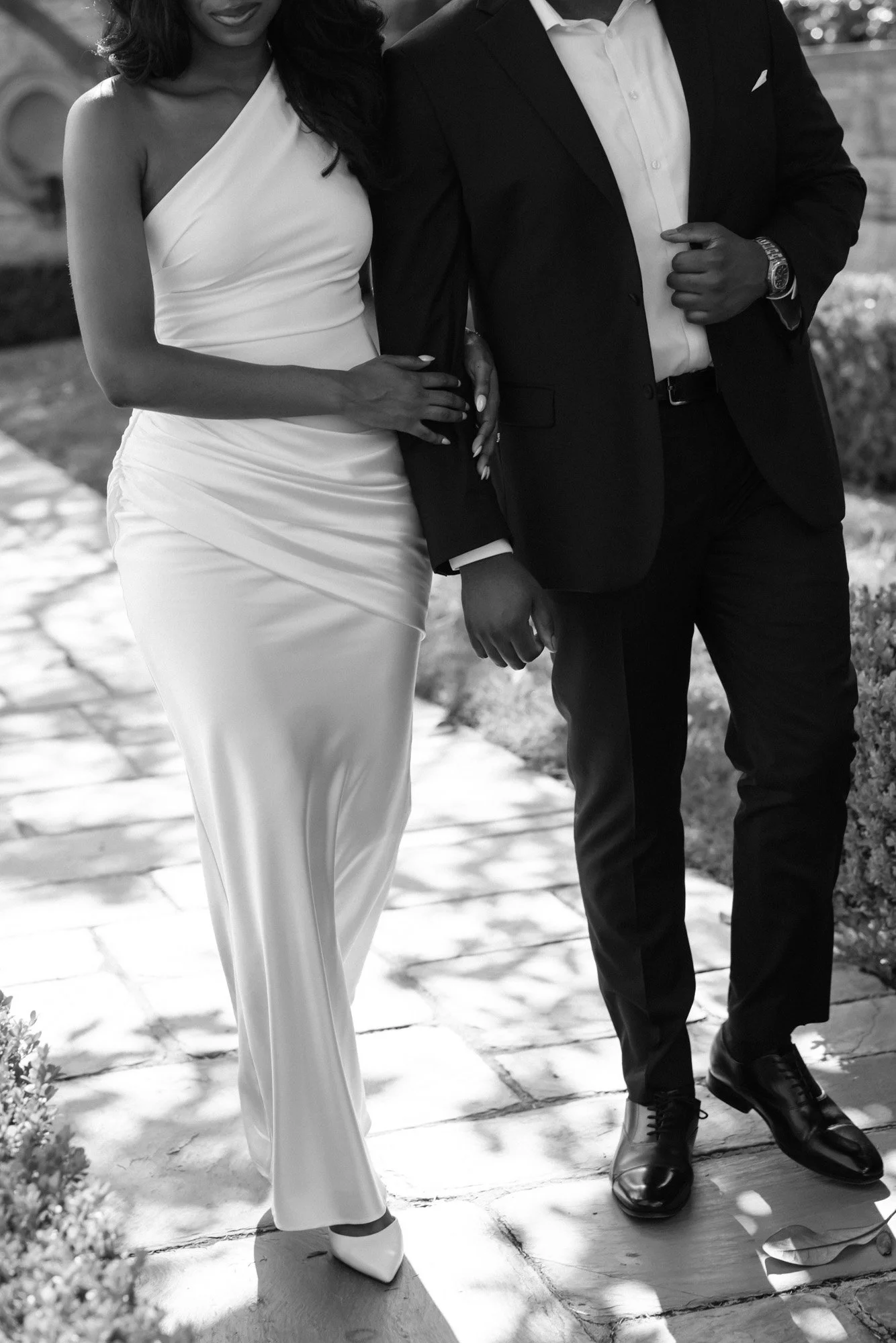 Black and white photo of a couple standing close together outdoors. The woman is wearing a one-shoulder white dress and high heels, and the man is in a dark suit with a white shirt and dark shoes. Both are holding hands.