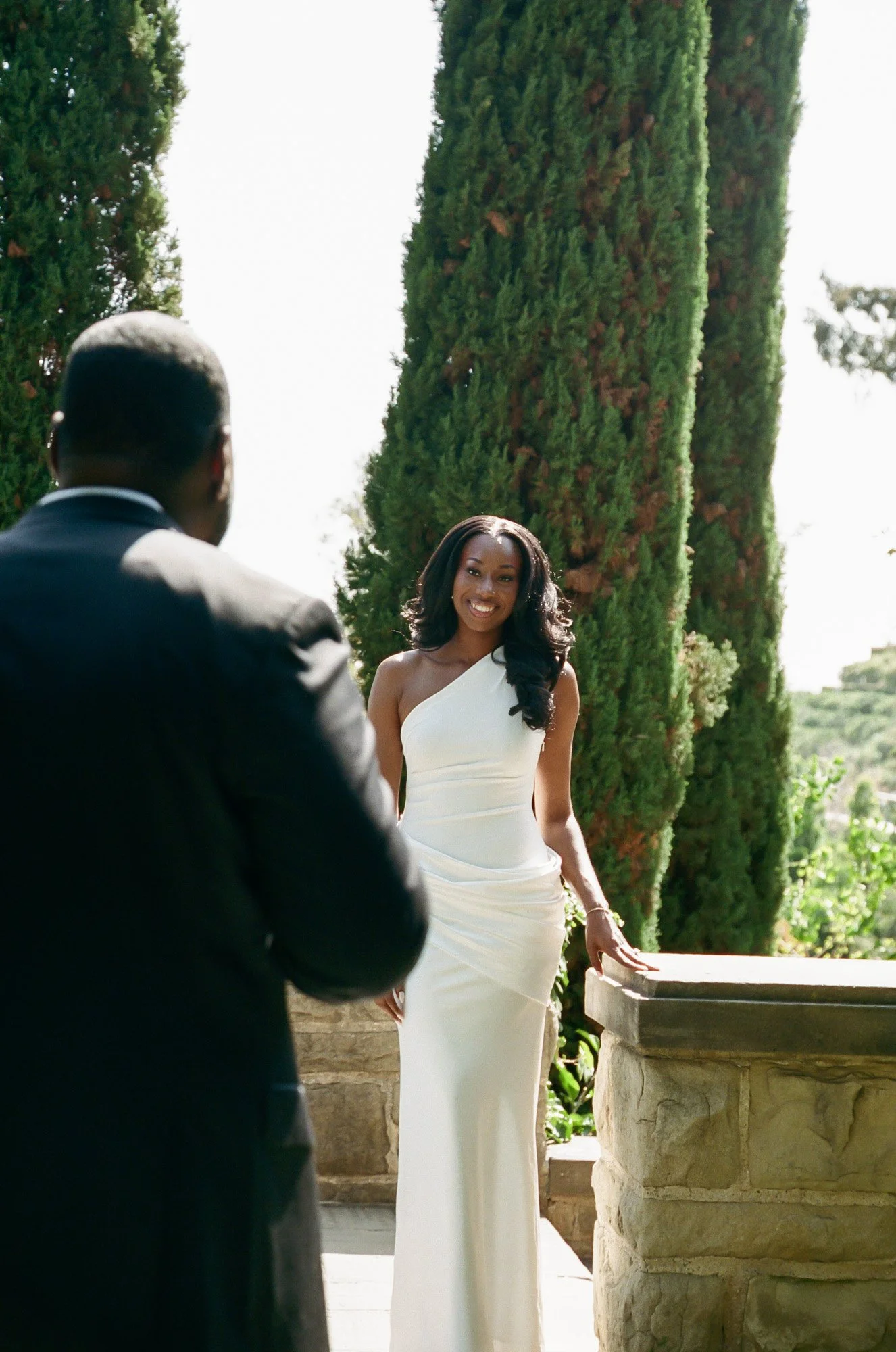 A woman in a white dress smiling at a man in a dark suit during an outdoor event with tall green trees in the background.