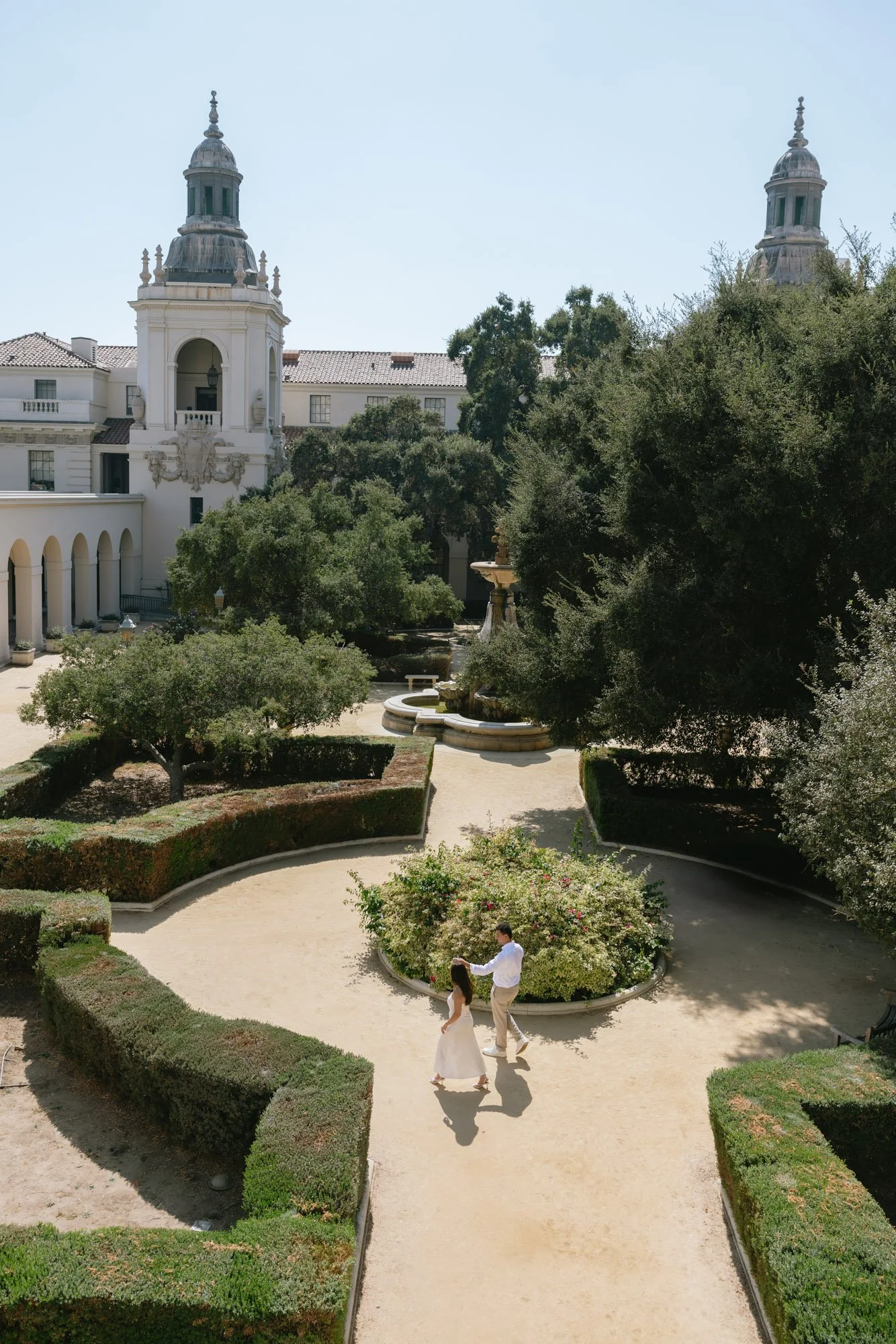 A man and woman dressed in wedding attire are holding hands and walking along a dirt path in a formal garden with hedges, trees, and a fountain. There are two large towers of a building with ornate architecture in the background. The sky is clear and