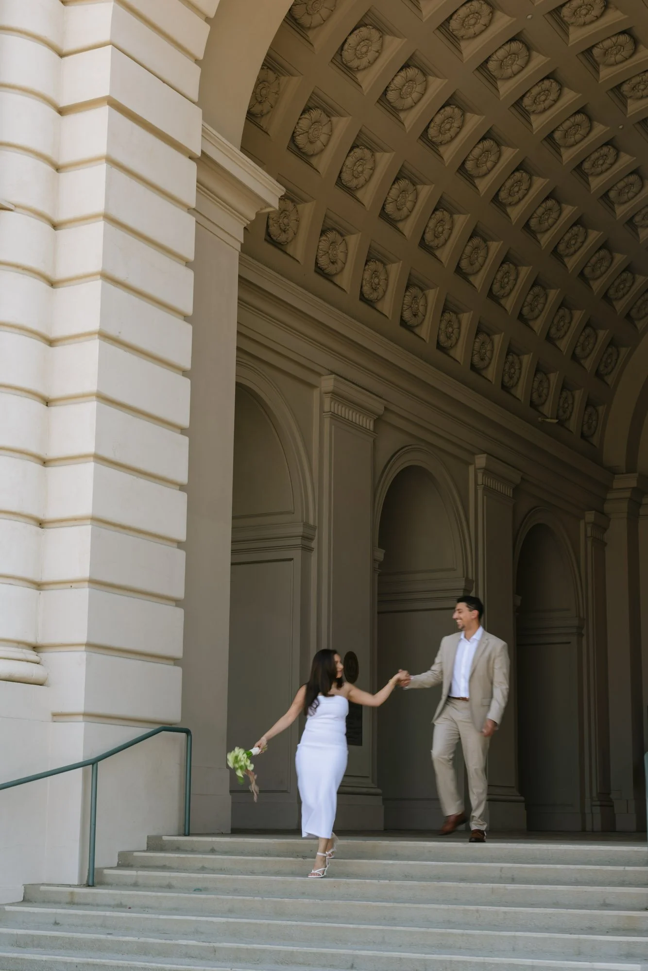 A couple dressed in formal attire on the steps of a building, with the woman holding a bouquet of flowers and the man extending his hand, smiling, under a decorative architectural ceiling.