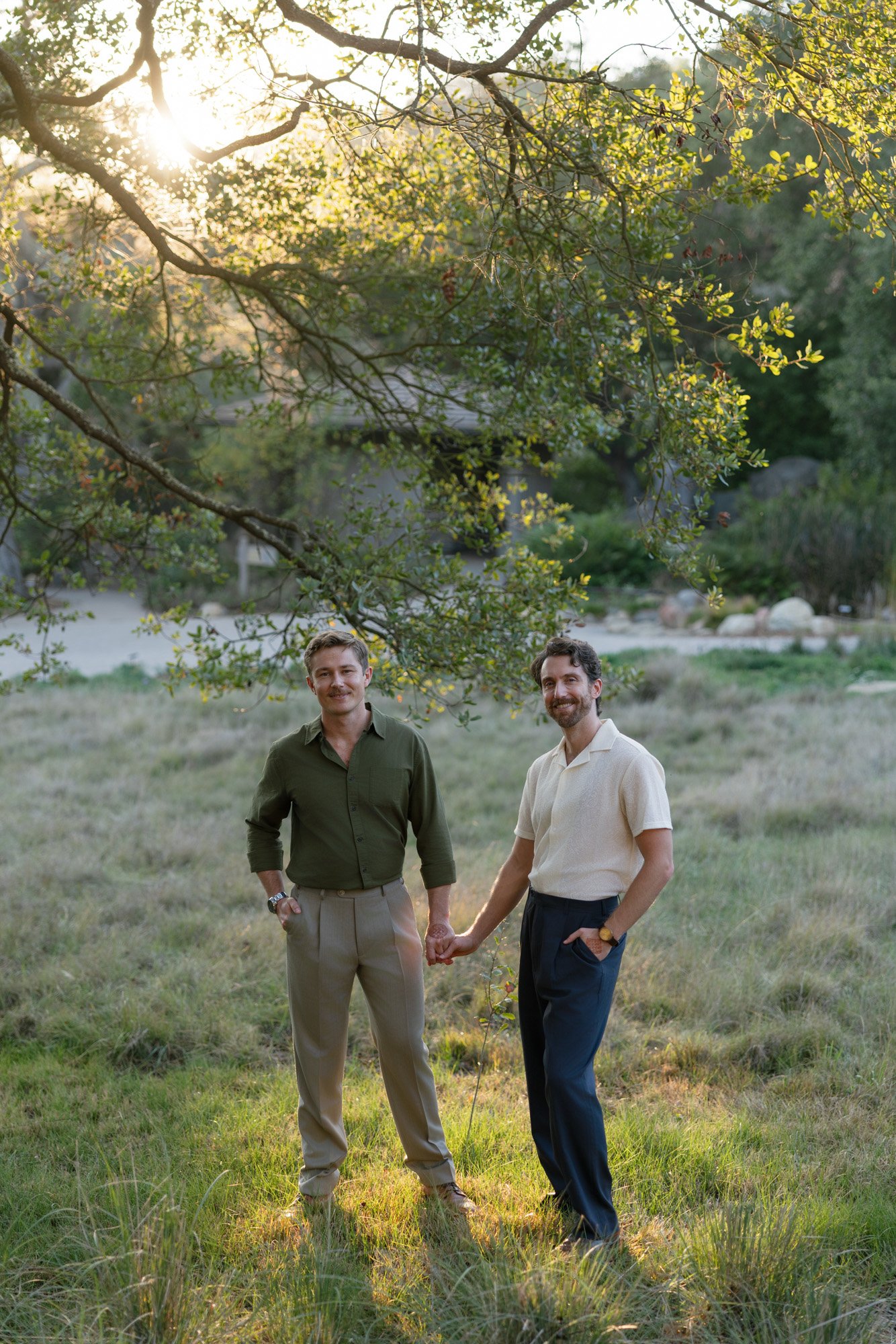 Two men holding hands outdoors in a grassy field with a large tree and a house in the background, during late afternoon or early evening.