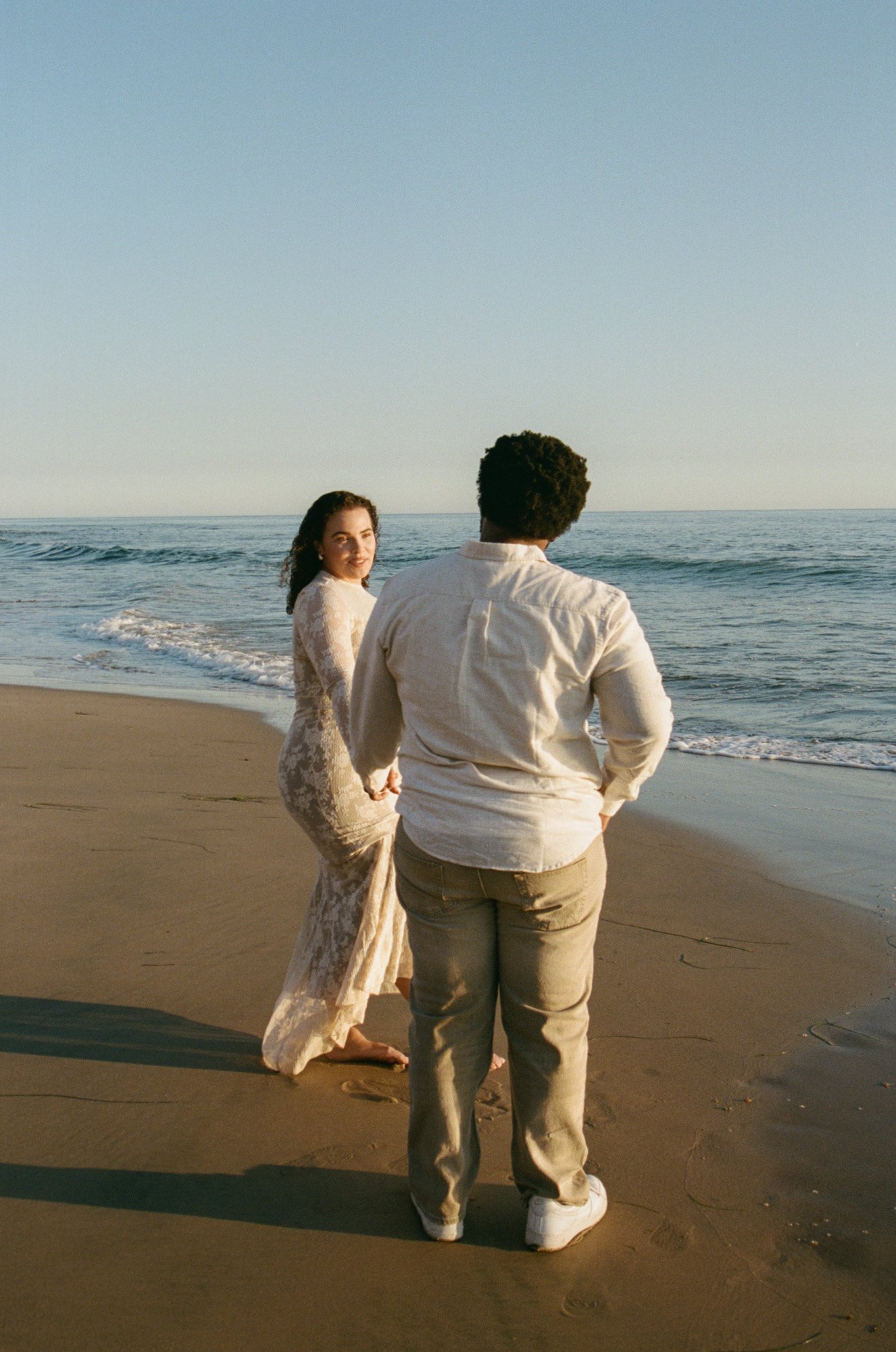 A couple stands on a beach, holding hands, with the ocean in the background during sunset. The woman wears a long, lace dress, and the man is dressed in casual khaki pants and a white shirt.