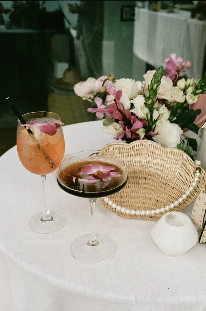 A table decorated with pink and white flowers, two cocktails with flower petals inside, a wicker purse with pearls, and a small white vase on a white tablecloth.