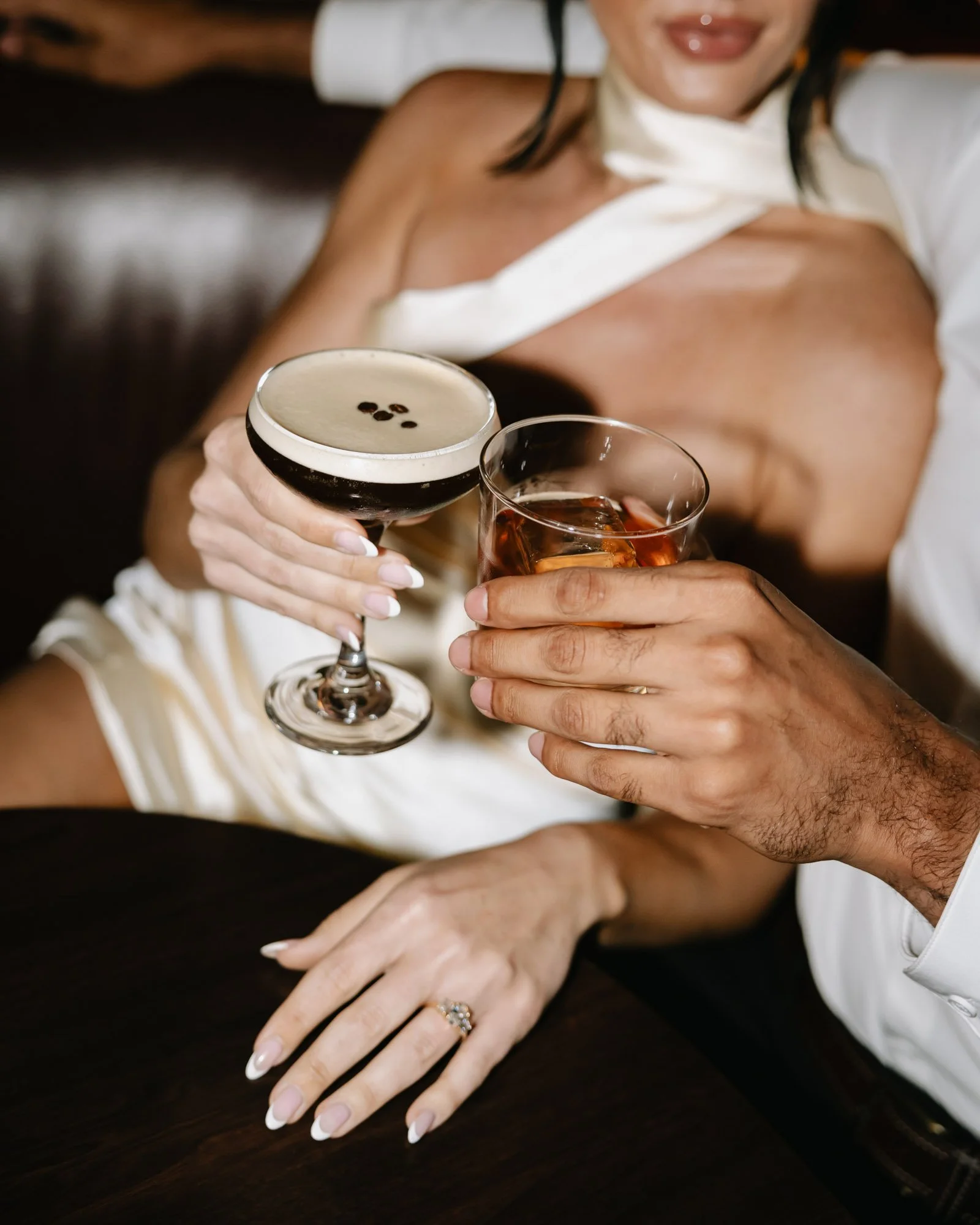 A woman is holding a glass of dark beer topped with foam and three coffee beans, while another person is holding a glass of amber-colored drink with ice, with their hands resting on a dark wooden surface.
