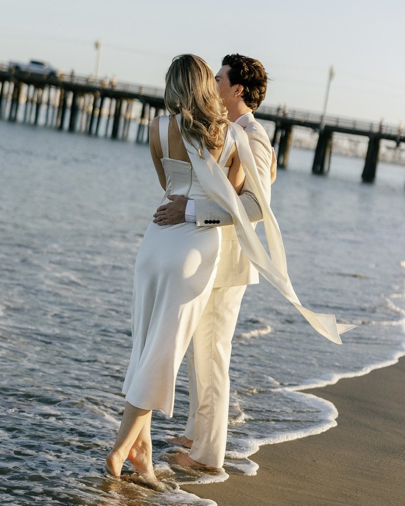 A couple in white clothing sharing a kiss at the beach near a pier at sunset.
