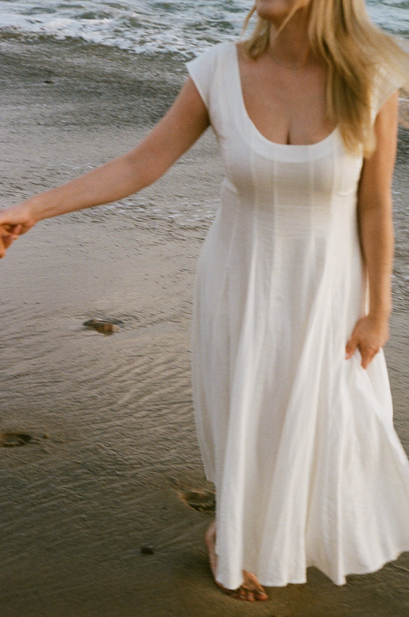 A woman in a white dress walking on the beach with bare feet, holding the hem of her dress, near the shoreline with wet sand and small rocks.