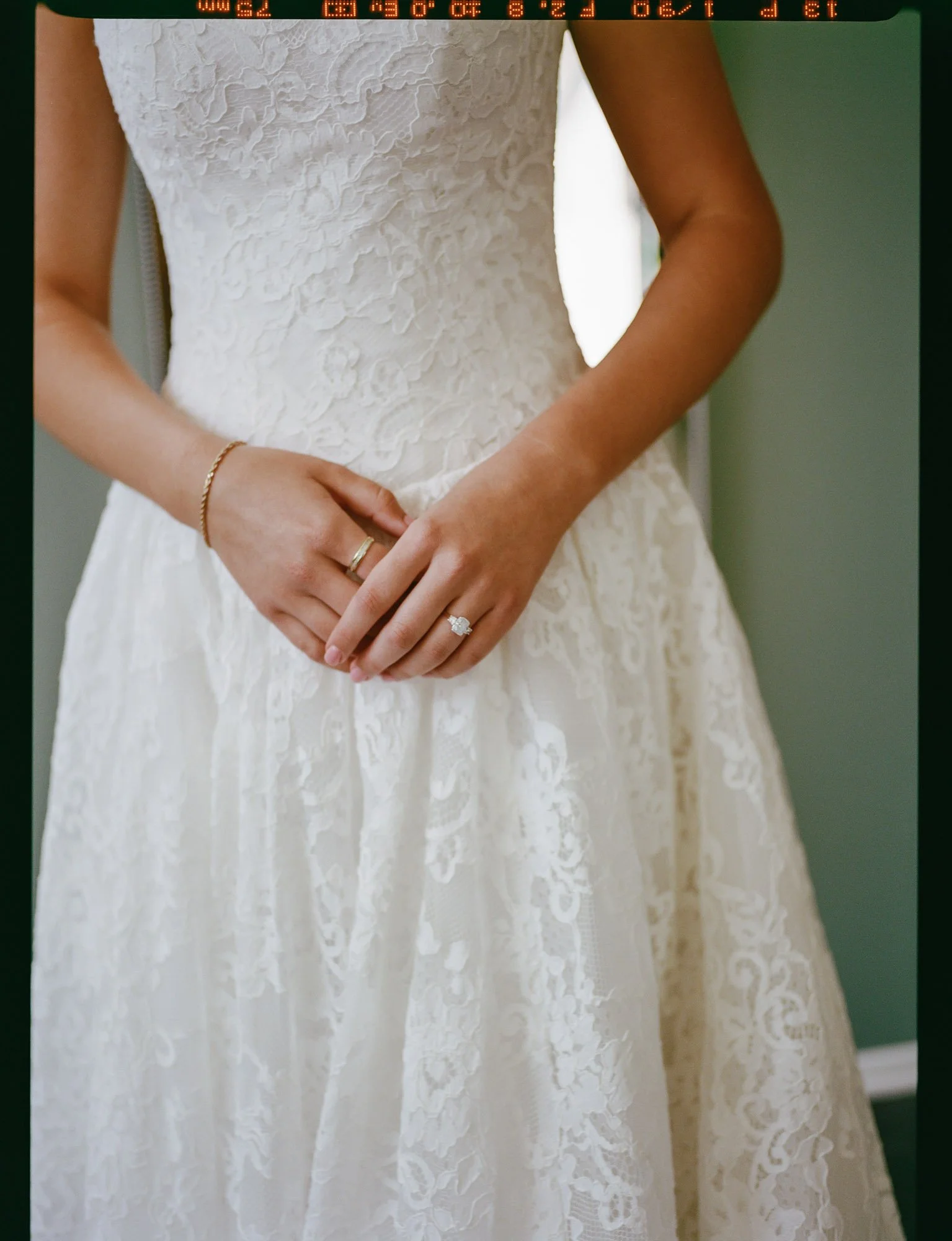 Close-up of a woman wearing a lace wedding dress, with hands clasped in front, showing an engagement and wedding ring, with a green wall in the background.