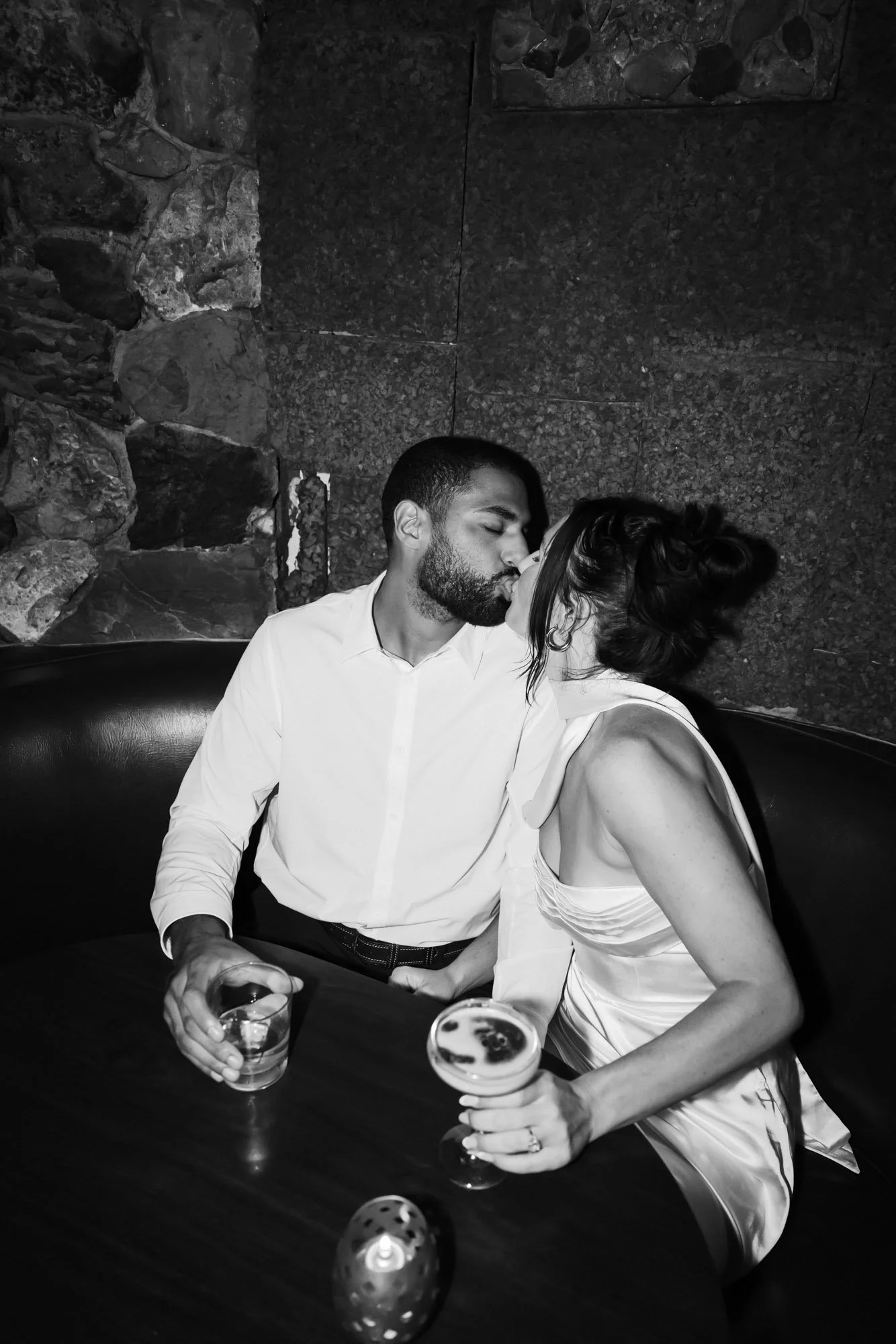 A black and white photo of a man and woman kissing while sitting at a table in a bar or restaurant. They are holding drinks, and some dice are on the table.