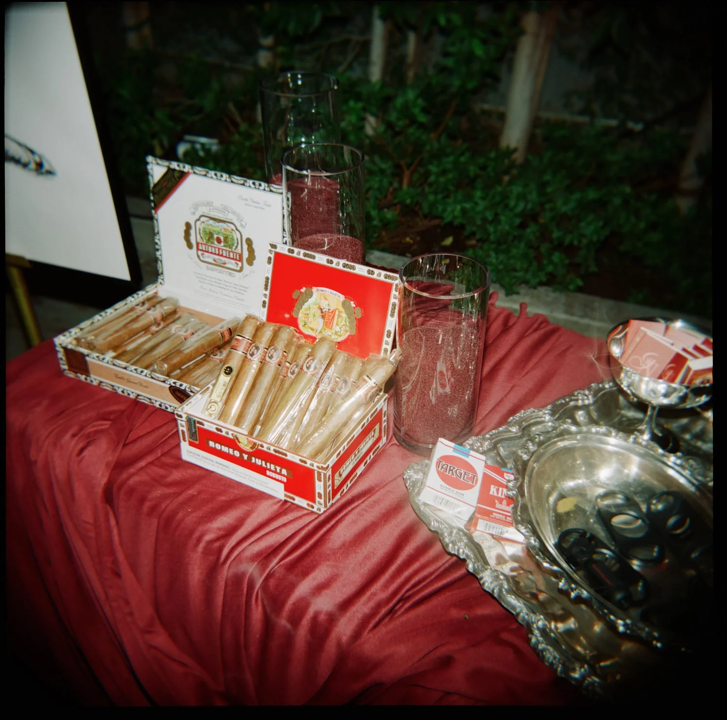 A display of cigars, cigar bands, and cigar-related items on a table with a red velvet cloth, including boxes of 'Romeo y Julieta' cigars, glass candle holders, and a silver tray with lighters.