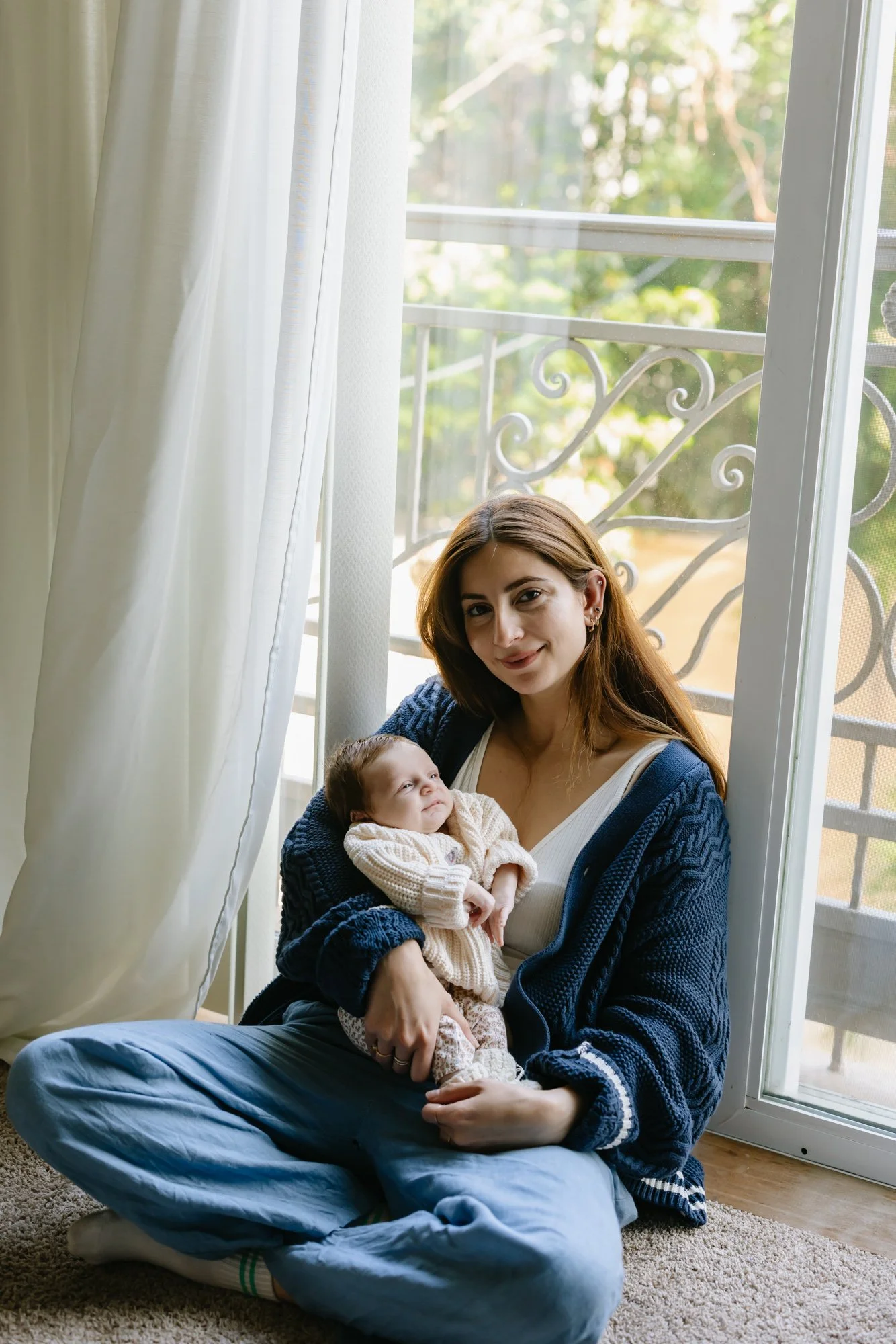 A woman sitting on a rug by a window, holding a baby in her lap, with natural light and greenery outside.
