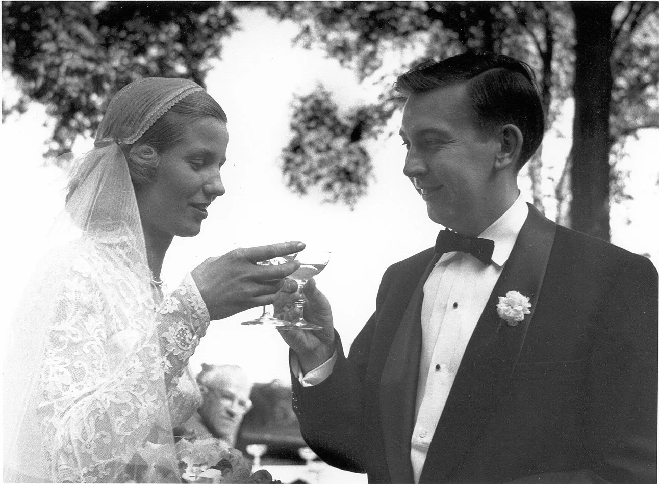 A black-and-white photograph of a wedding ceremony outdoors, featuring a bride in a lace dress and veil, and a groom in a tuxedo with a bow tie and boutonniere, clinking glasses.