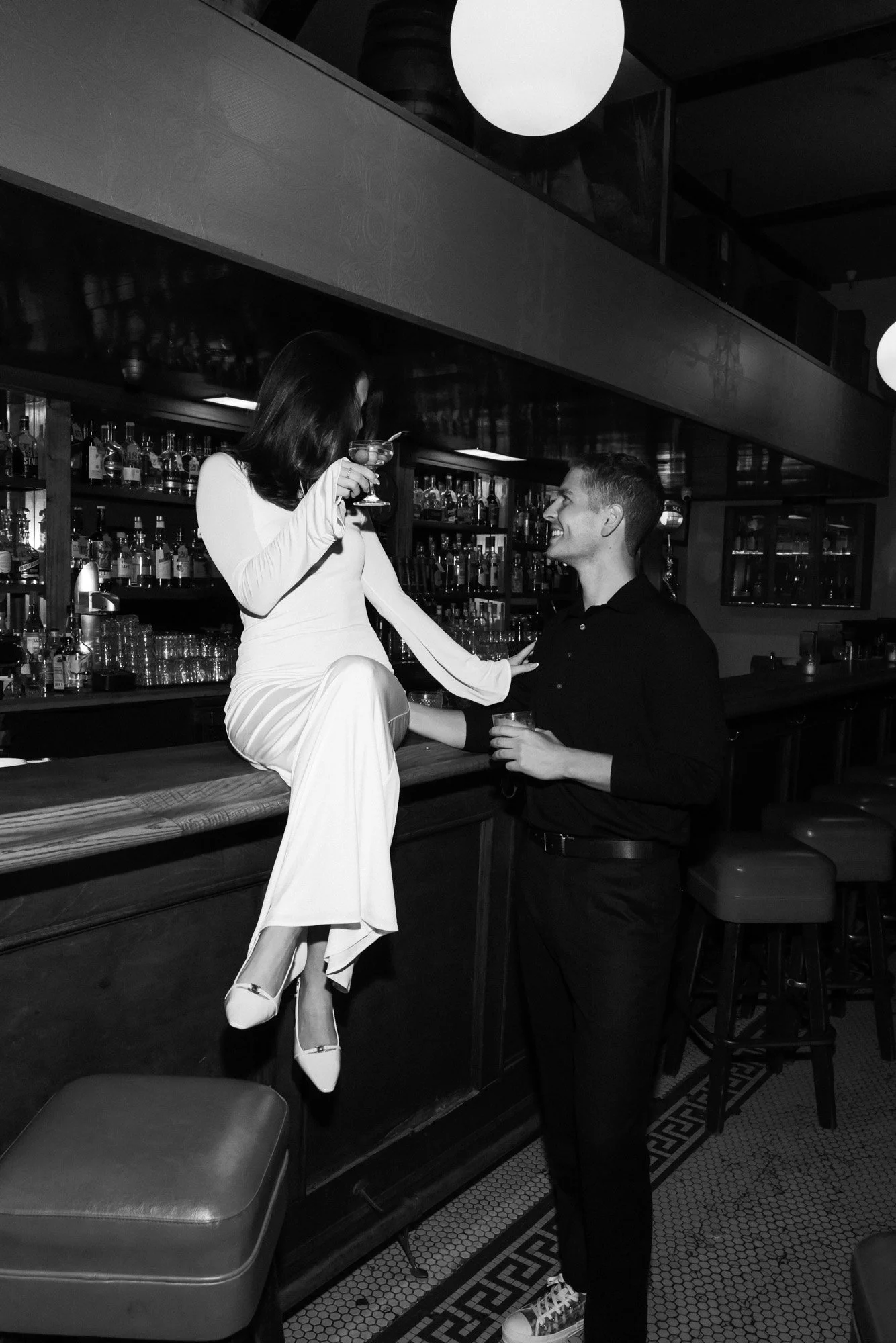 A woman in a white dress sitting on a bar, holding a cocktail glass, talking to a man in black shirt and pants behind the bar in a dimly lit restaurant or bar.