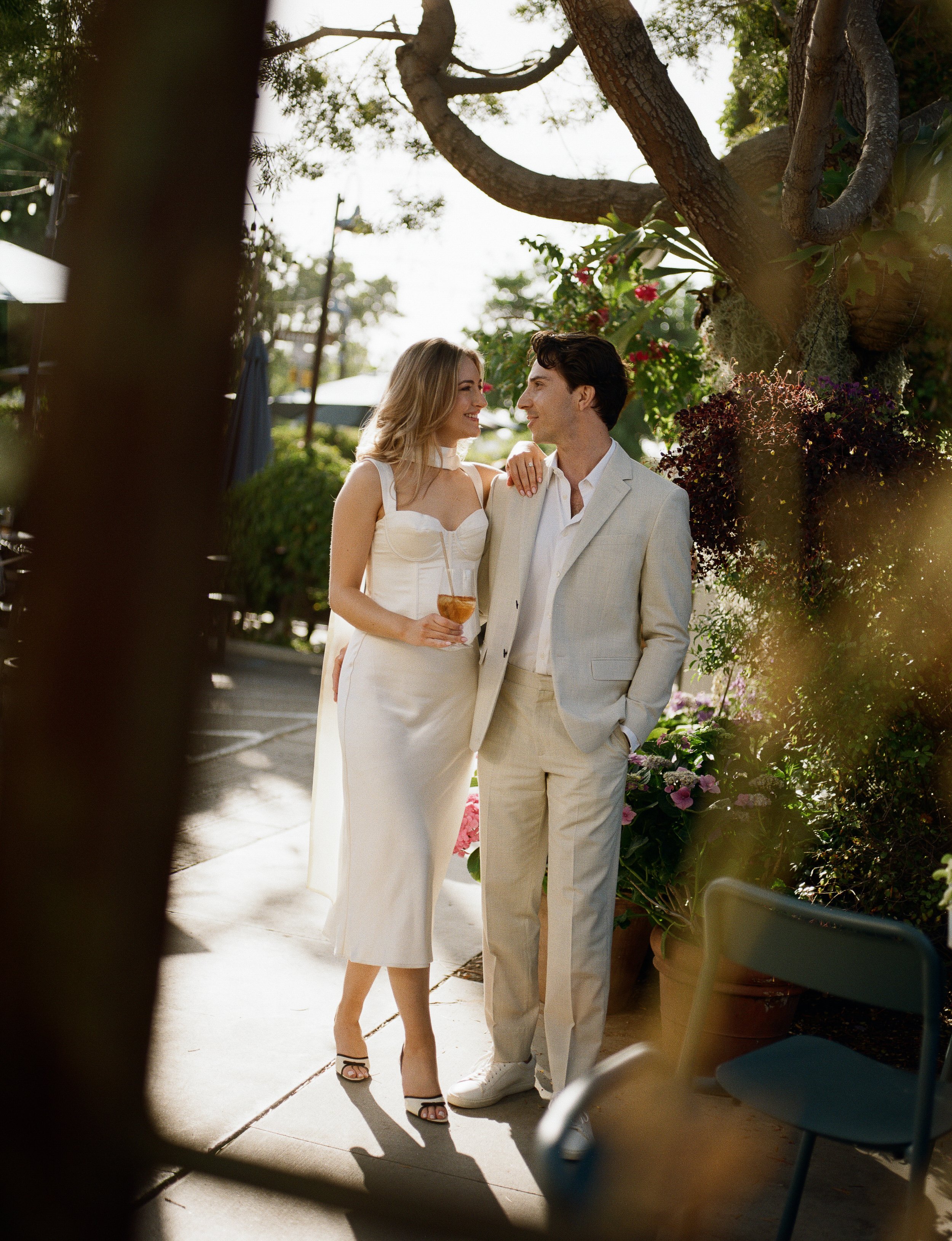 A couple standing close together outdoors, smiling at each other, dressed in wedding attire, surrounded by greenery and potted plants.