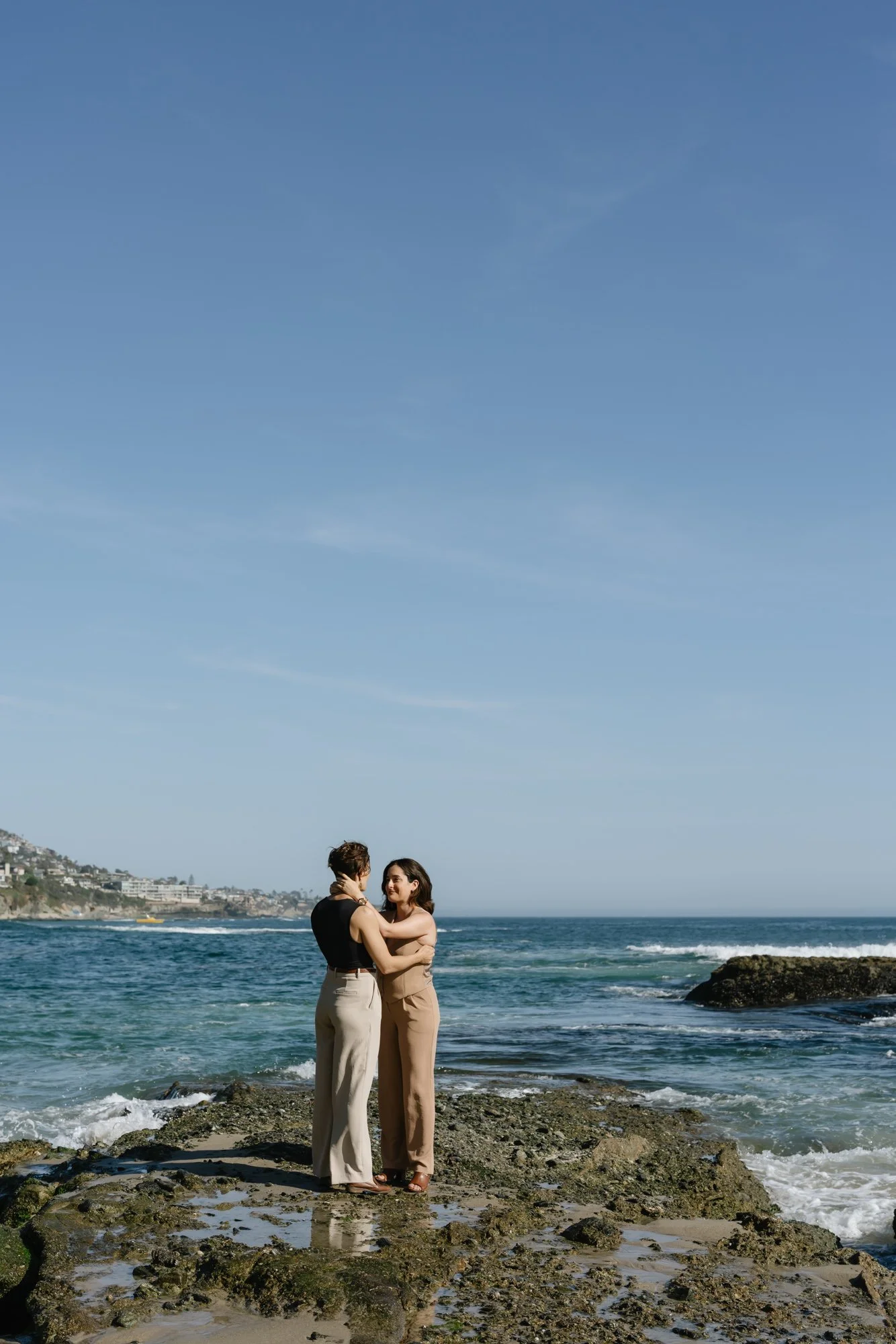 Two women standing on rocks at the beach, embracing each other with the ocean and a hilly coastline in the background under a clear blue sky.