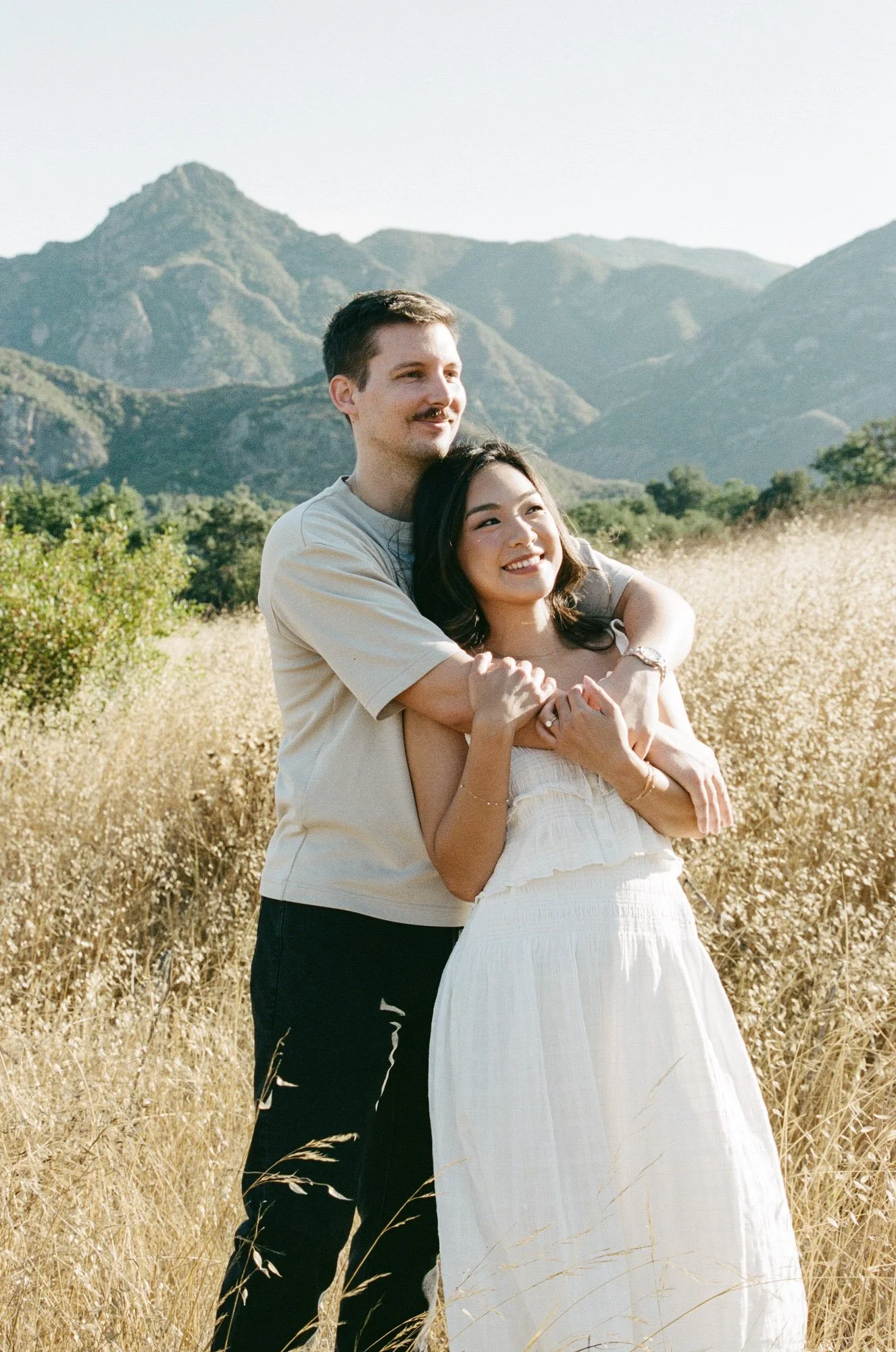 A man and woman sharing a joyful hug in a golden field with mountains in the background, sunny weather.