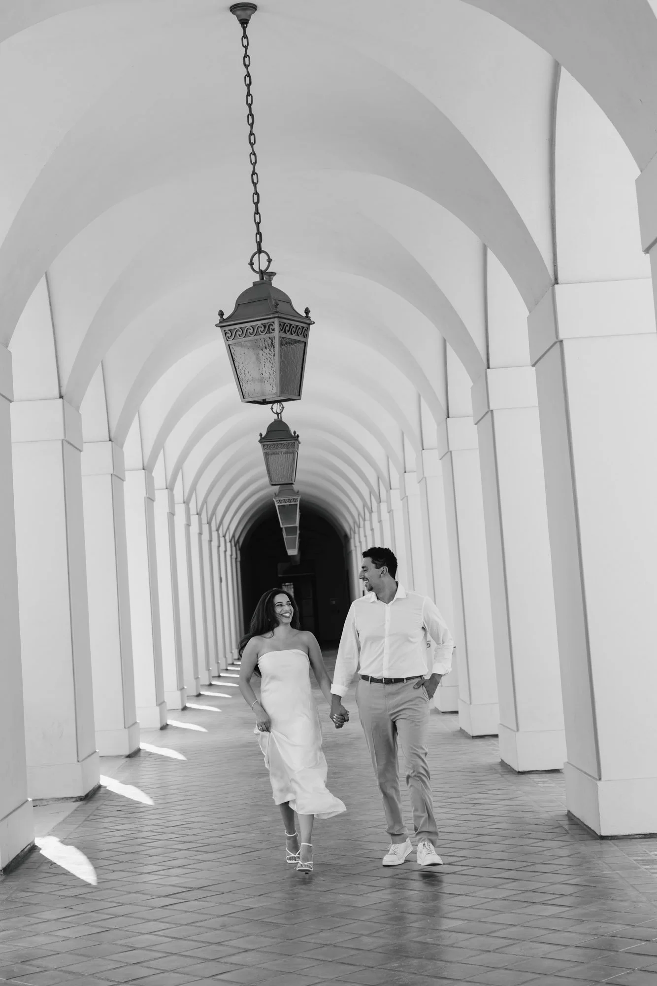 A smiling couple walking hand in hand through a covered walkway with white arches and hanging lanterns, in black and white.