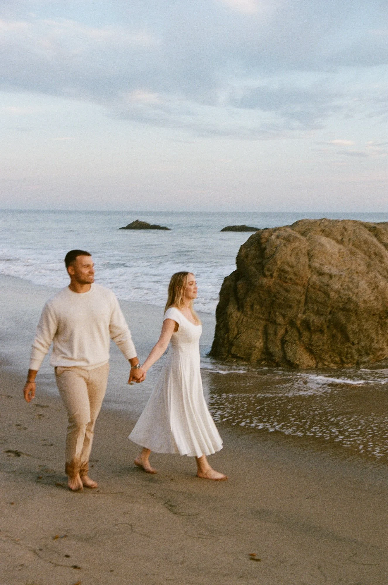 A couple dressed in light-colored clothing walking hand-in-hand along a sandy beach near the ocean, with rocks and cloudy sky in the background.