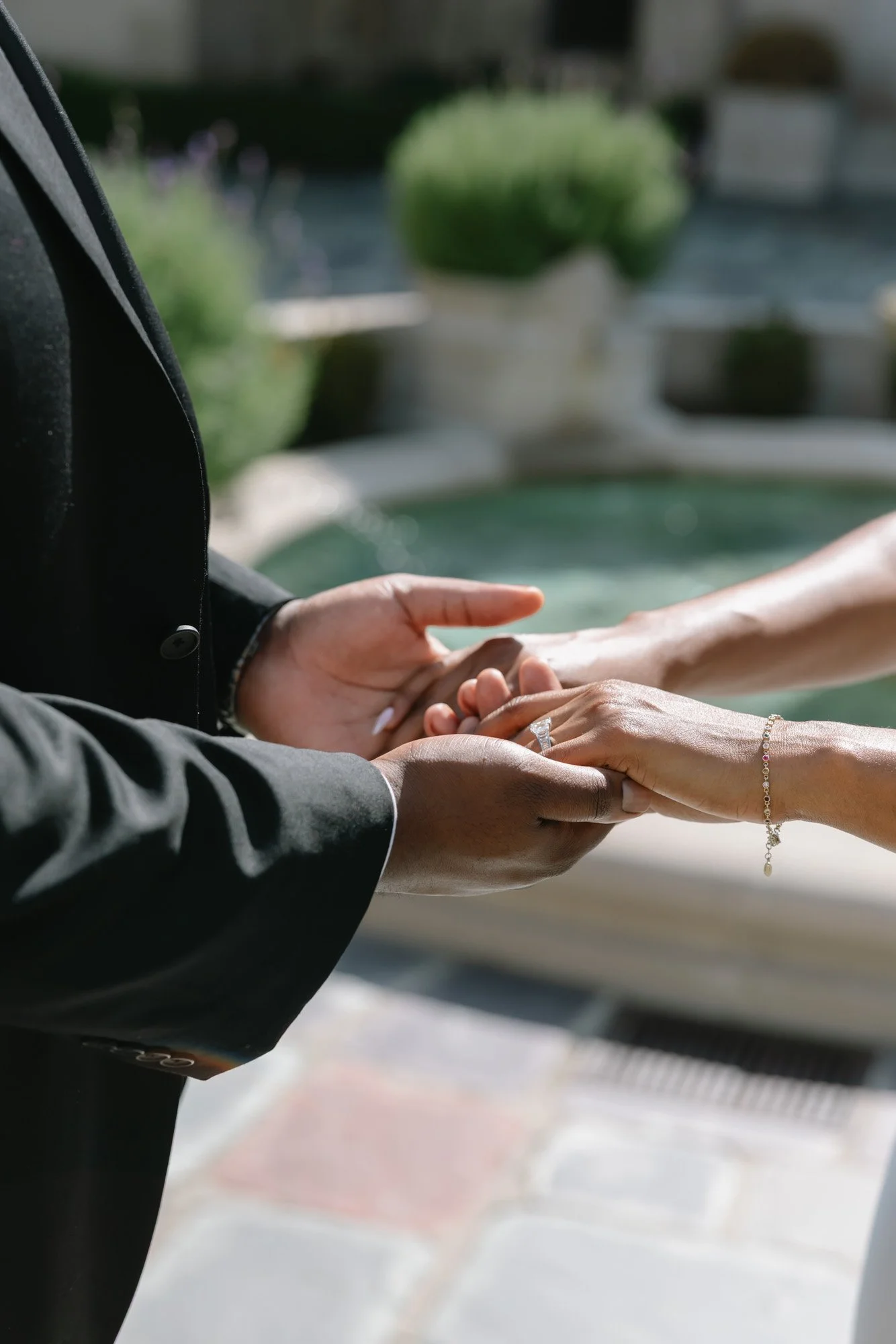 An engaged couple holding hands, with the man's hand gently touching the woman's hand, showing a wedding ring and bracelet, set outdoors with blurred green plants and a building in the background.