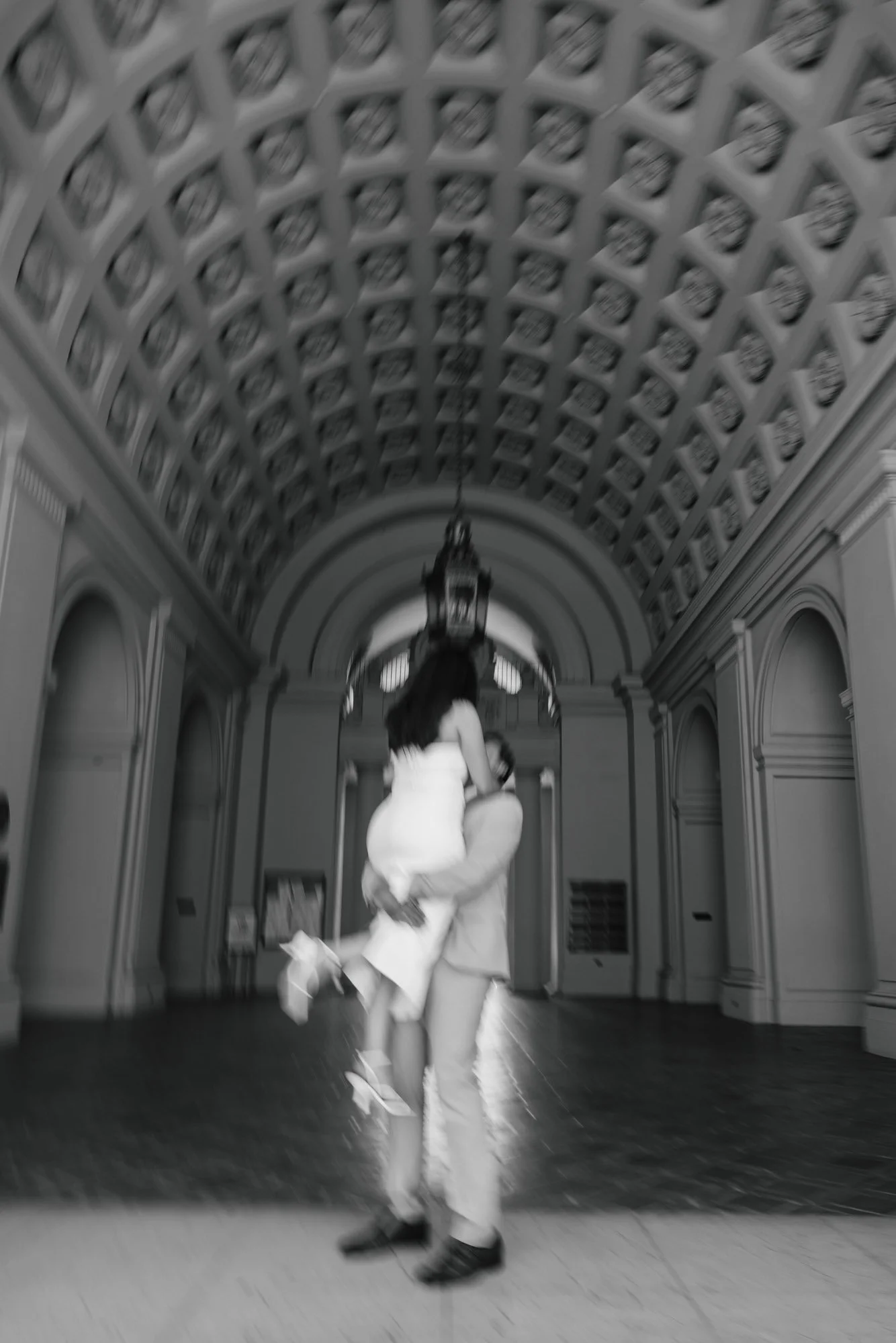 A black and white photo of a couple dancing in a grand hallway with a decorative ceiling and chandelier.