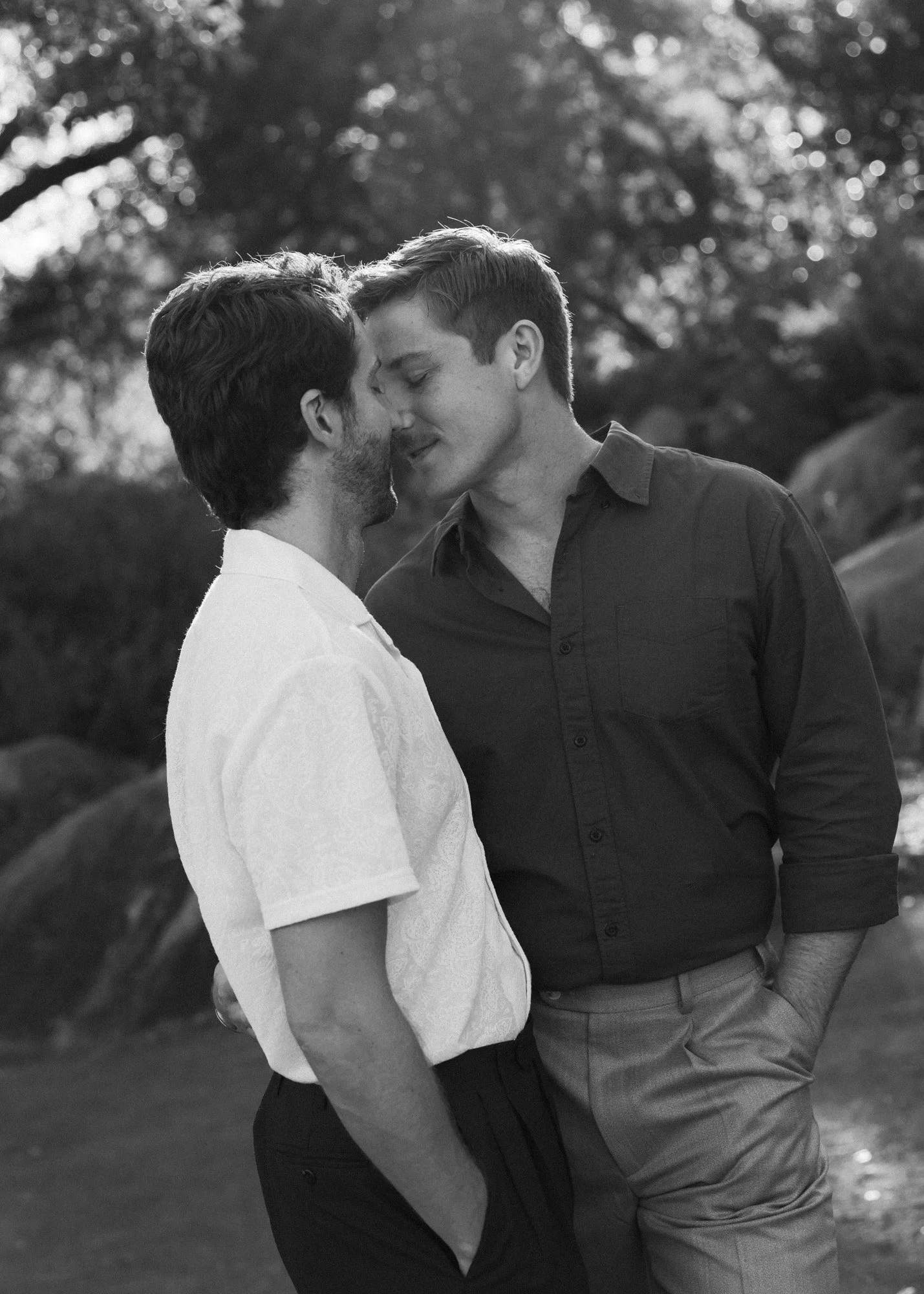 Black and white photo of two men standing close with foreheads touching, eyes closed, in a tender moment outdoors with trees in the background.