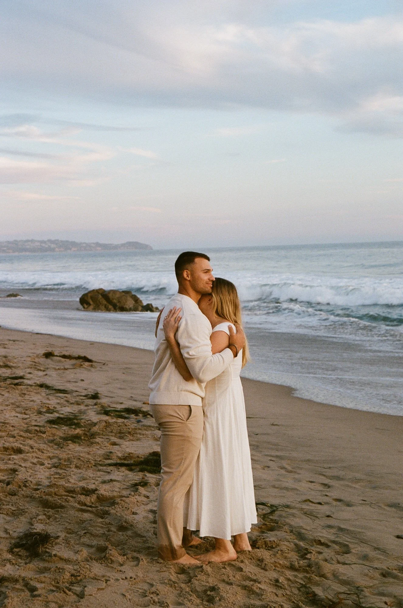 A young couple hugging on a sandy beach with ocean waves in the background, during sunset or dusk.