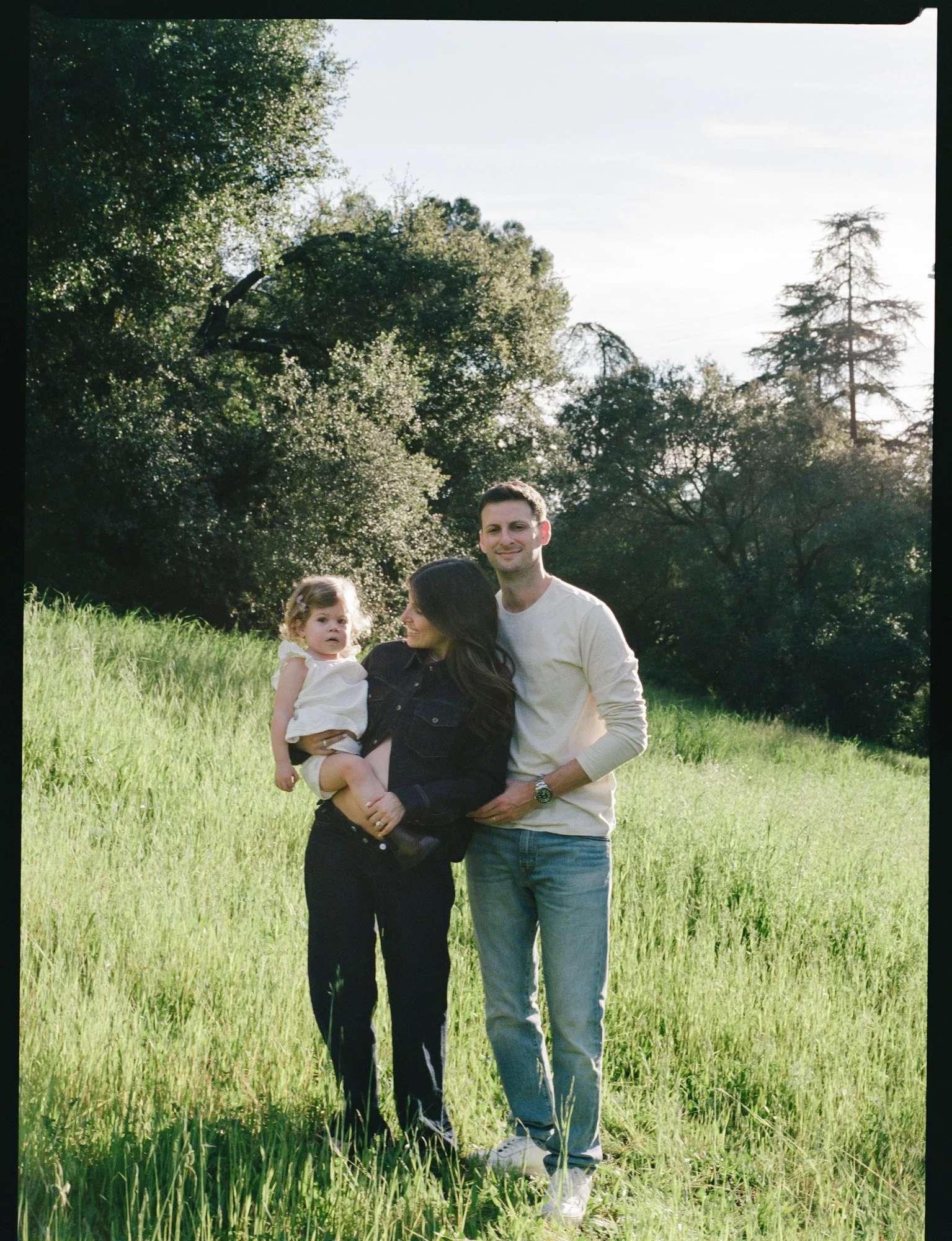 A family of three, including a woman, a man, and a young girl, standing outdoors in a grassy field with trees in the background, during daylight.