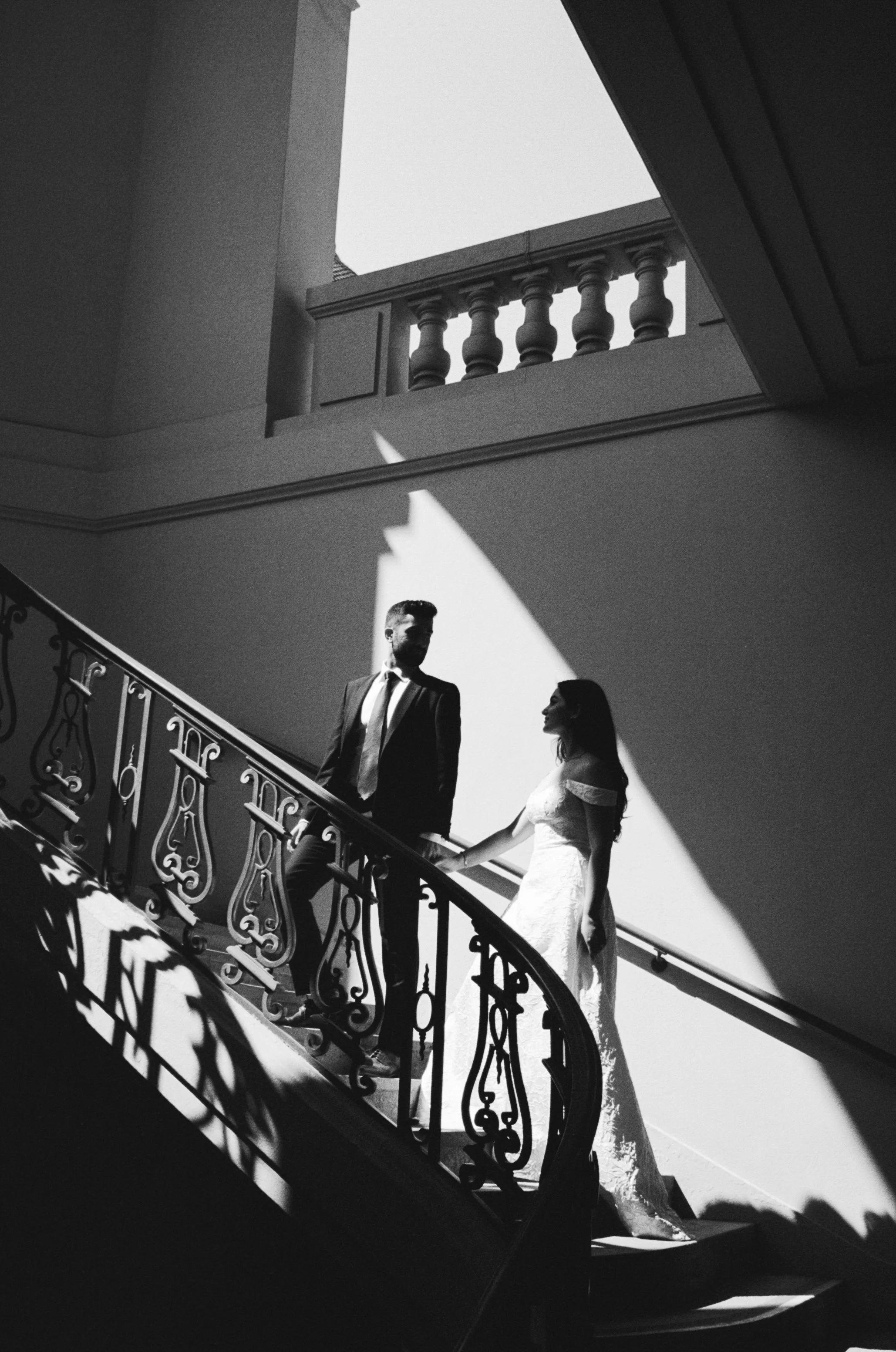 A black and white photograph of a bride and groom standing on a staircase. The bride in a wedding dress holds hands with the groom in a suit and tie. The staircase has ornate black wrought-iron railings, and sunlight casts shadows across the scene. T