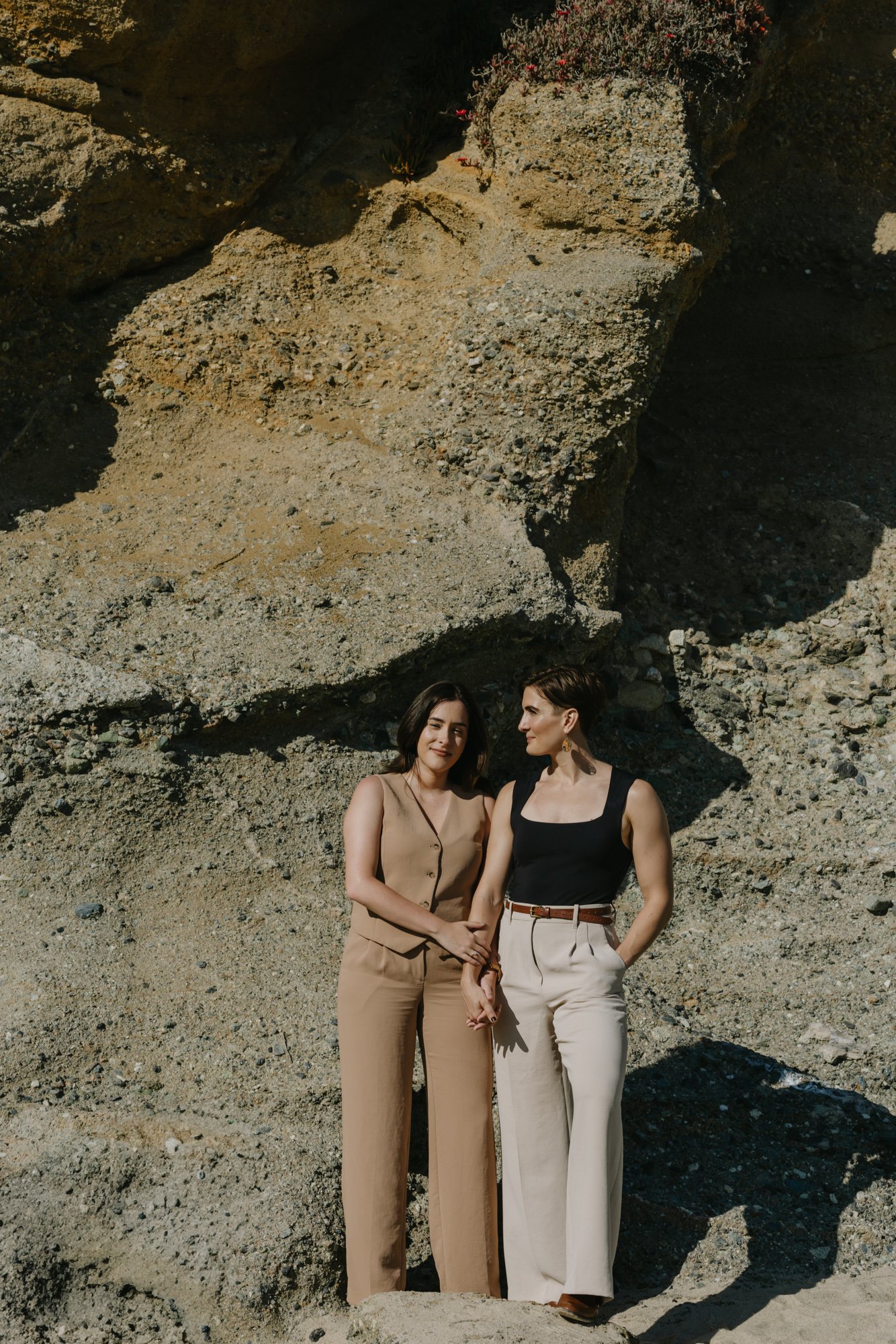 Two women stand together outdoors against a rocky terrain and large boulder. One woman has short dark hair, wearing a beige sleeveless vest and matching wide-leg pants. The other woman has short hair, wearing a black sleeveless top and light-colored 