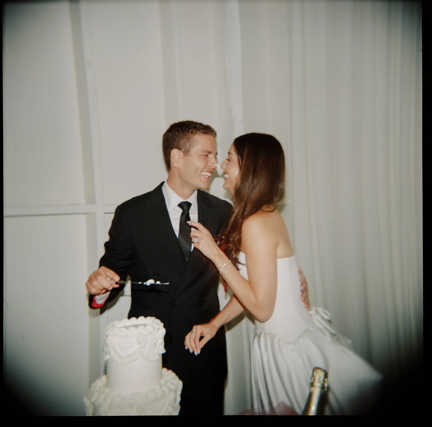 A newlywed couple at a wedding reception, smiling and looking at each other near a wedding cake.
