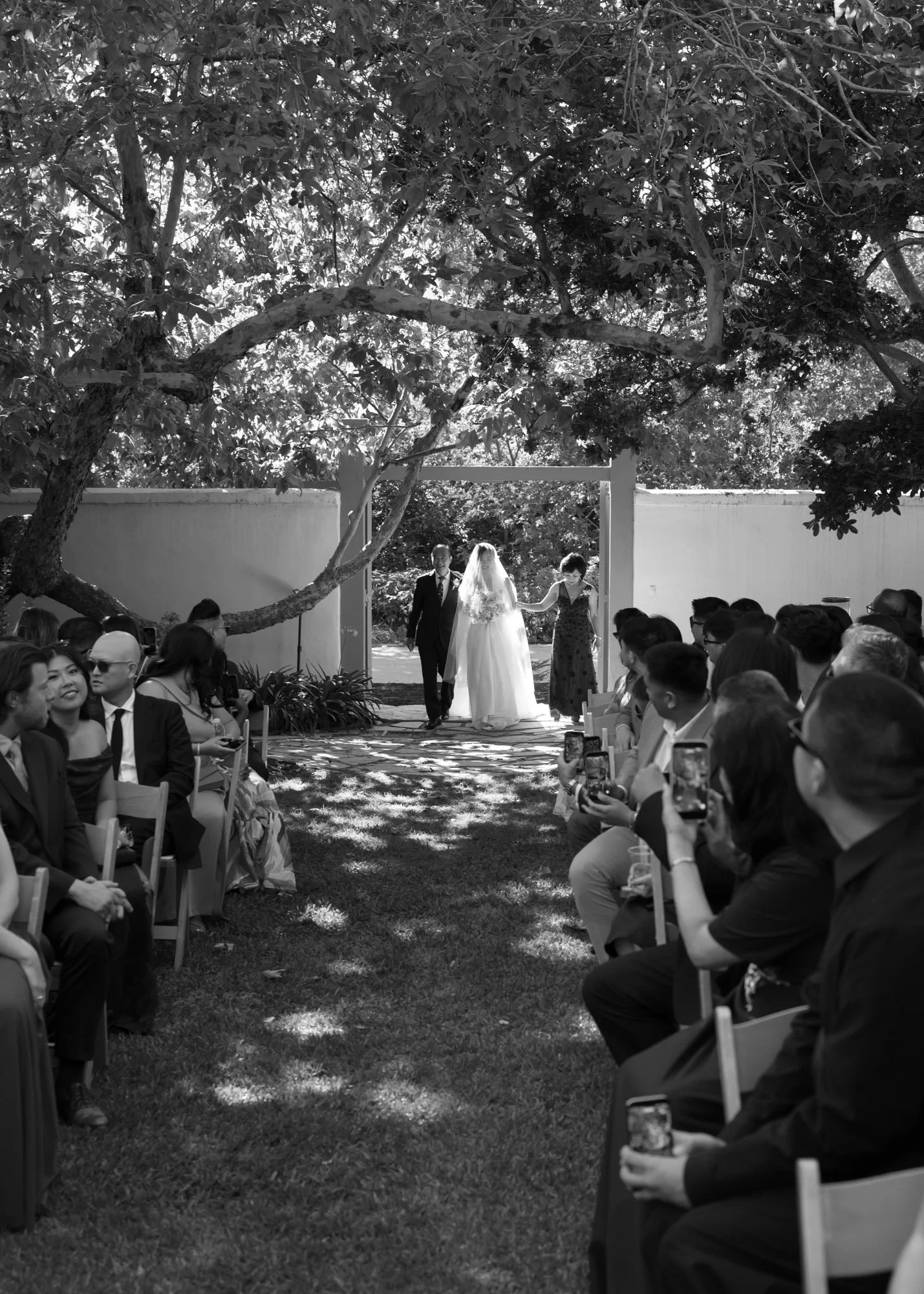 Black and white outdoor wedding ceremony beneath trees photographed in a documentary style
