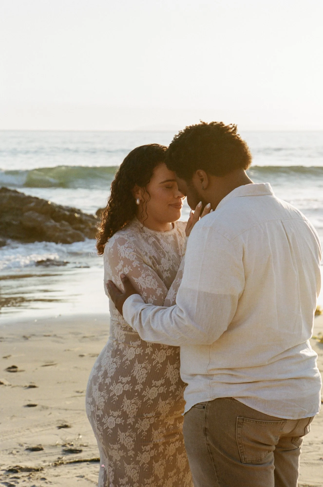 A couple dressed in white standing close on a beach at sunset, with the ocean and rocks in the background.