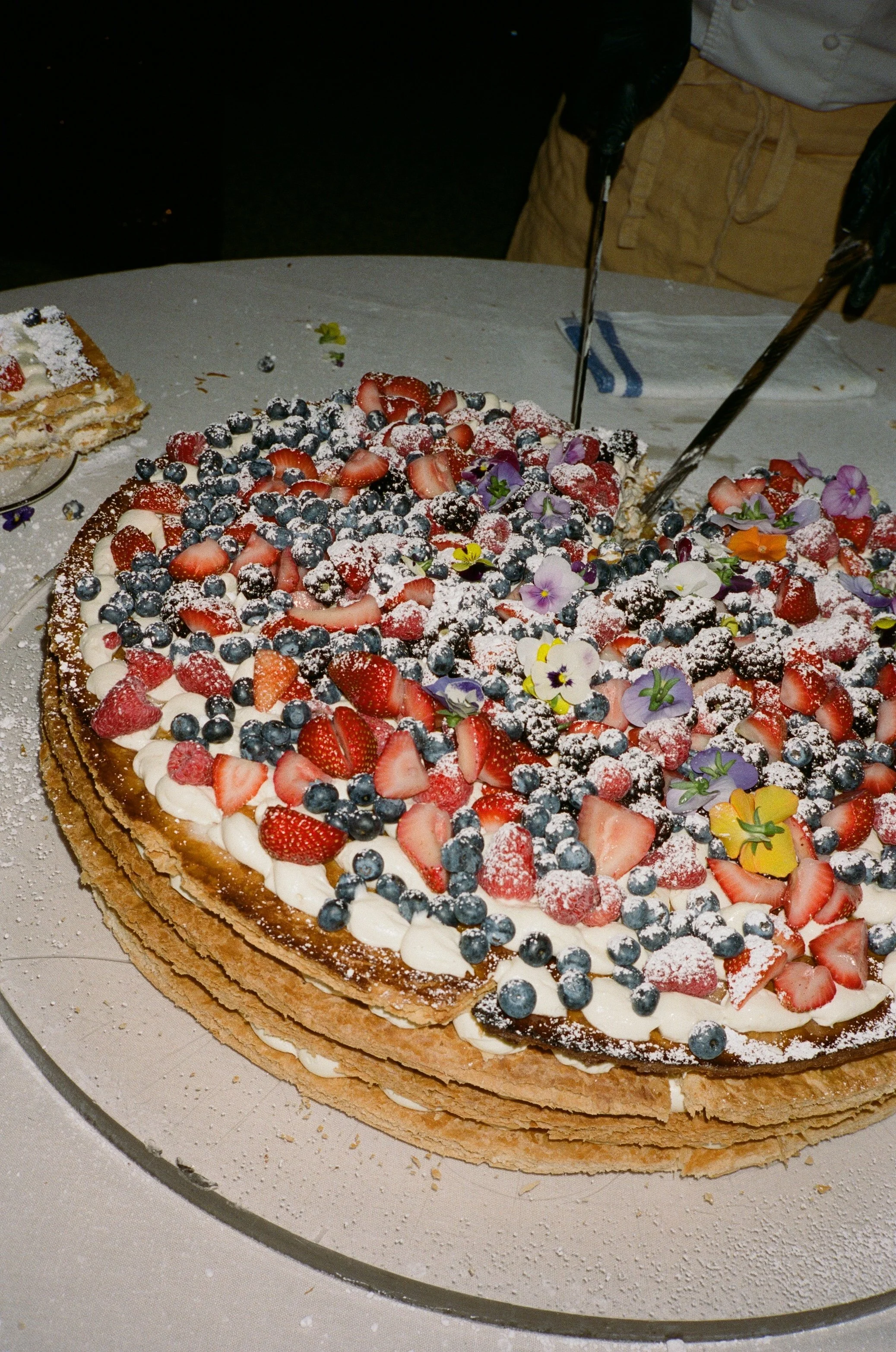 A large oval layered fruit cake topped with strawberries, blueberries, and edible flowers, dusted with powdered sugar.