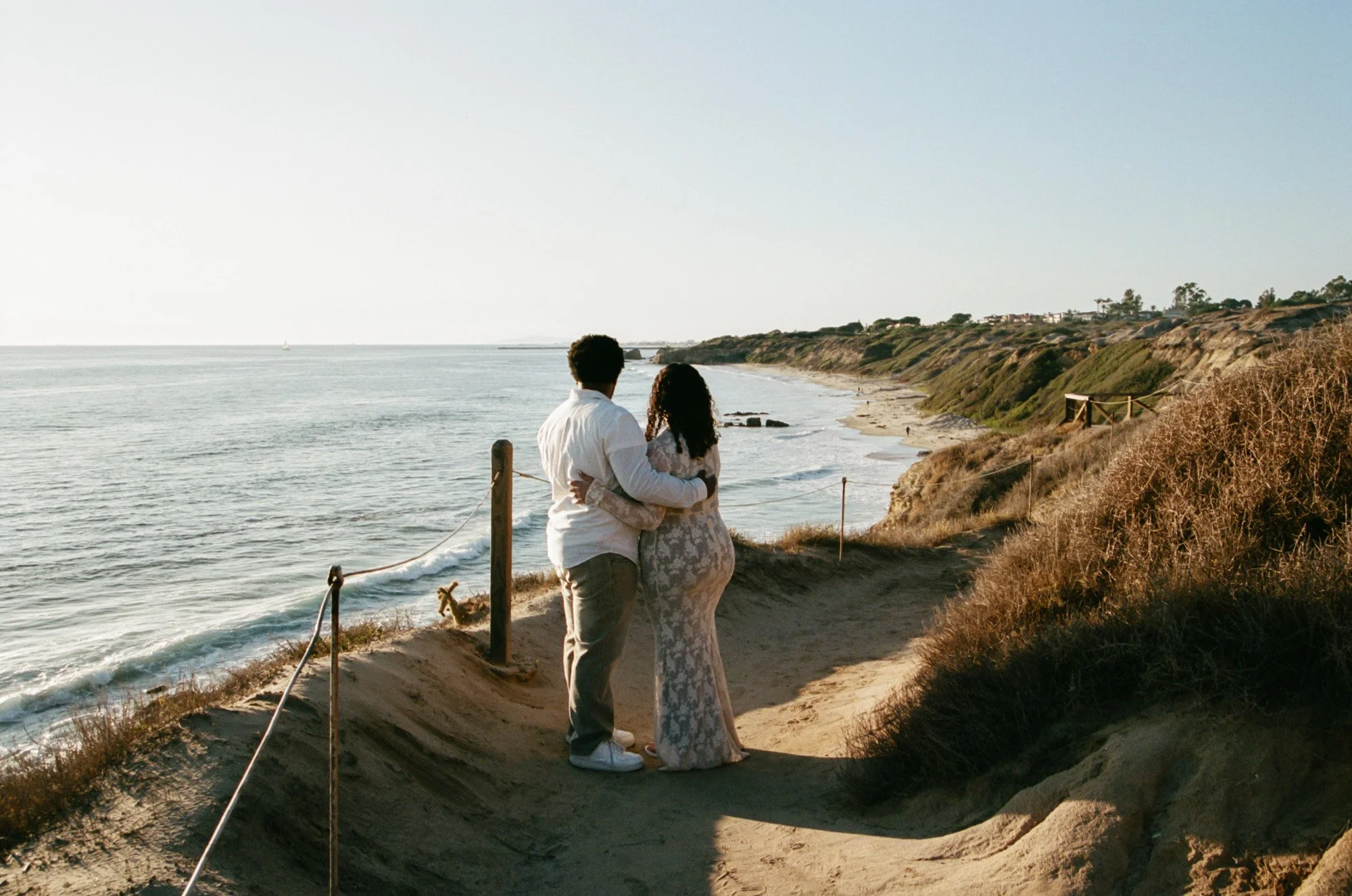 A couple stands on a sandy path near the ocean, with waves crashing on the shore, a cliff with houses in the background, and the sky clear and bright.