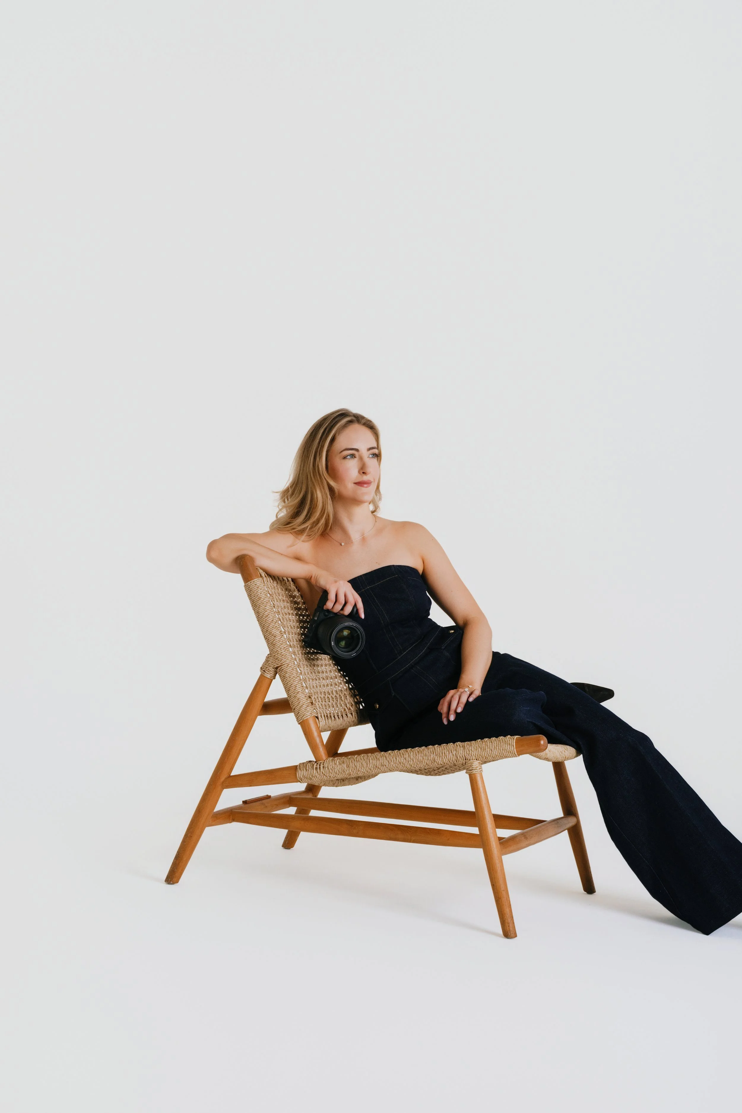 A young woman with blonde hair sitting on a wooden chair with a woven seat, holding a camera, and looking off to the side, against a plain white background.