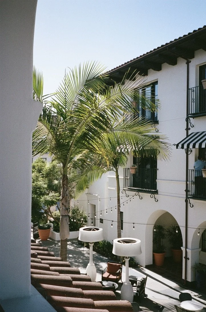A courtyard with tall palm trees, white walls, black window railings, string lights, and outdoor furniture, including two white patio heaters and a lounge chair.