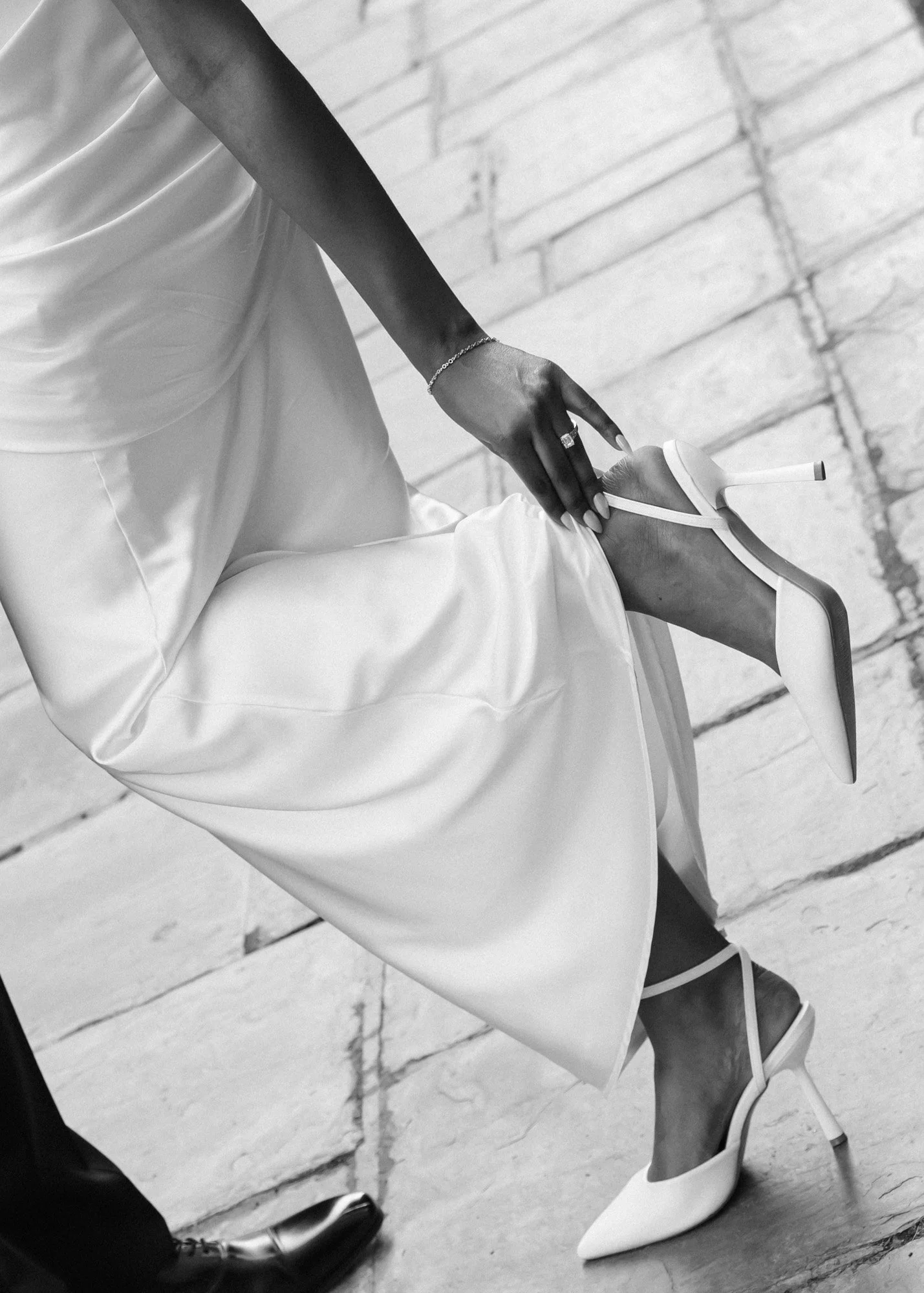 Close-up of woman’s legs and feet wearing white high heels and a satin dress, sitting on a chair, with a man’s black dress shoes and pant leg visible beside her. The floor is made of stone or concrete tiles.