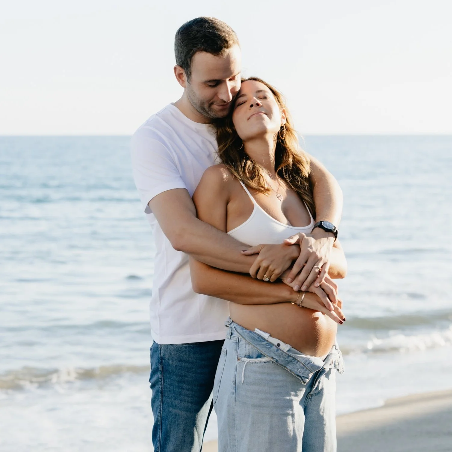 A couple hugging by the ocean, with the man embracing the pregnant woman from behind, both with closed eyes and content expressions.