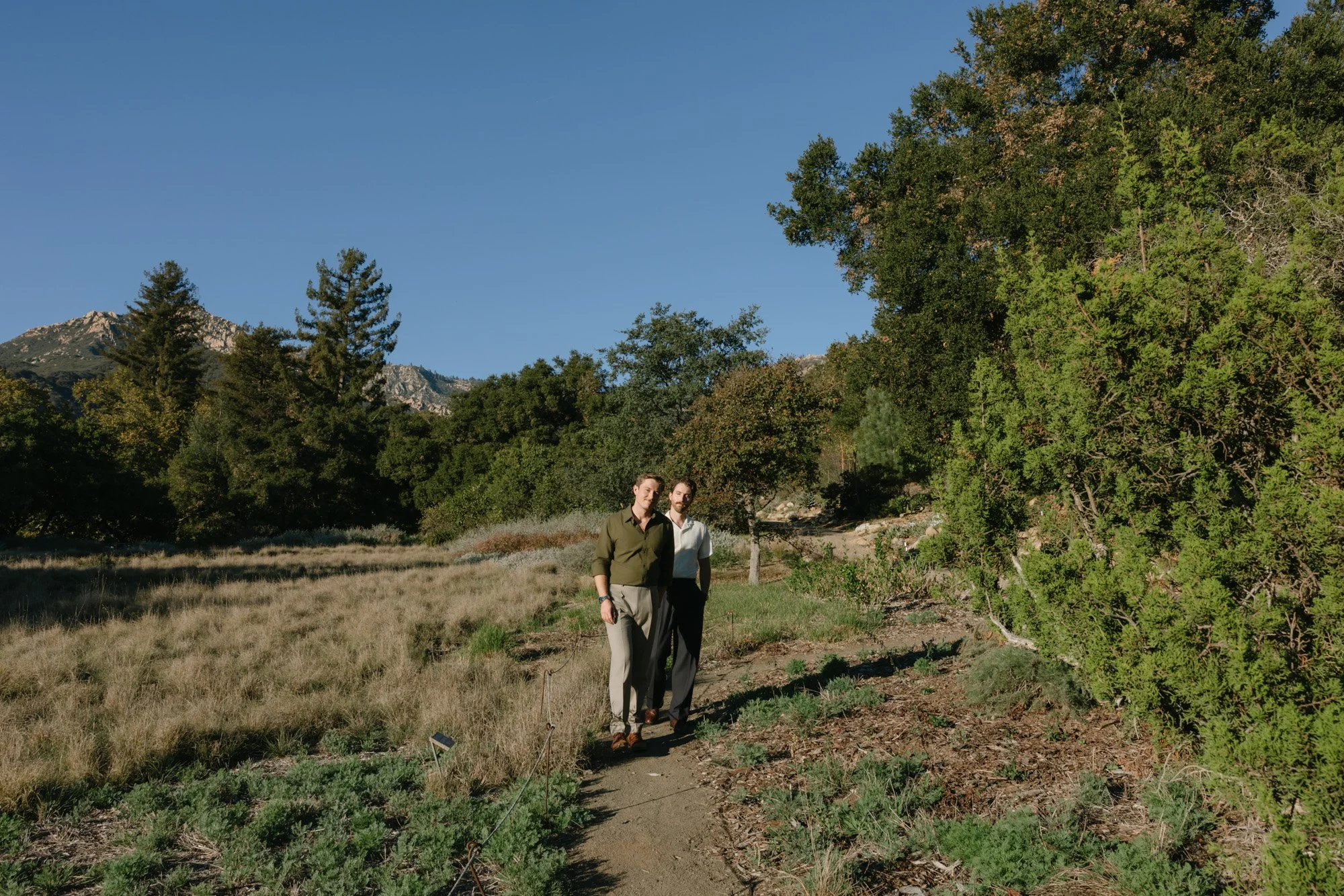 Two men walking on a dirt path through a grassy field surrounded by tall trees and mountains under a clear blue sky.