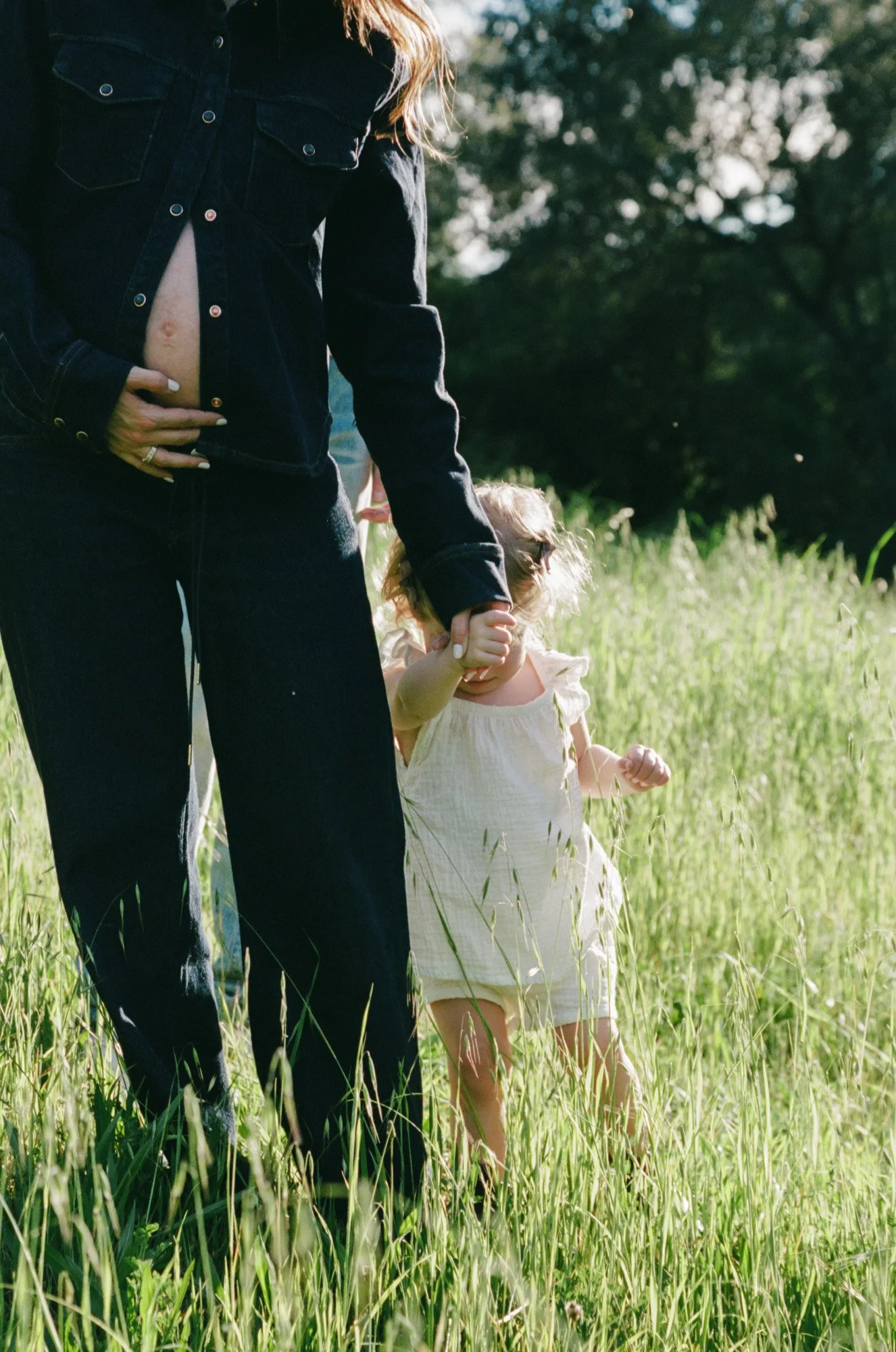A woman holding a young girl by the hand in a grassy field with trees in the background.