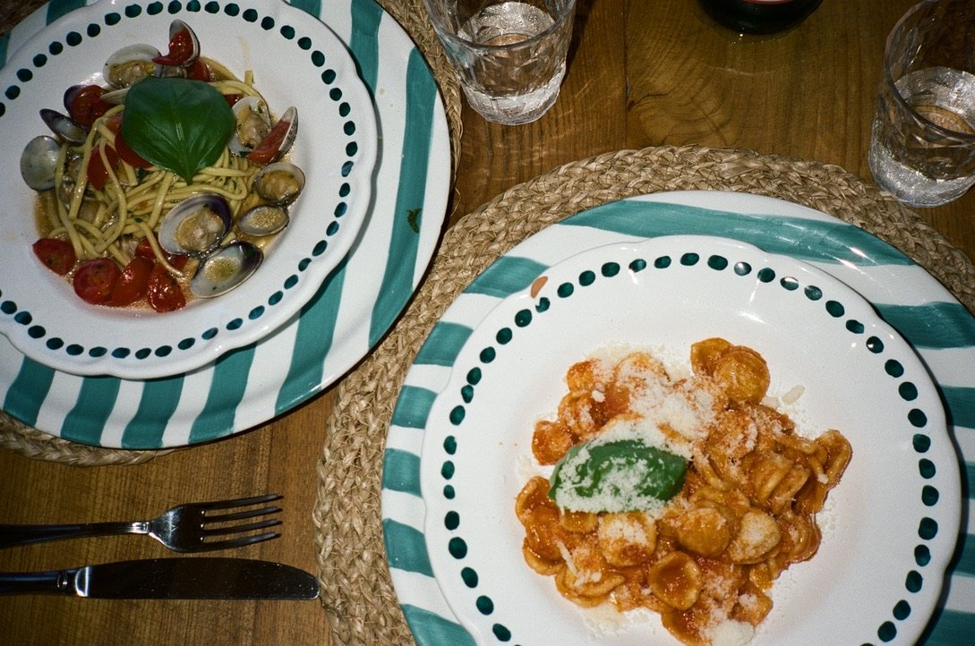 A table with two plates of food: one with pasta and cherry tomatoes topped with a basil leaf, and the other with gnocchi in tomato sauce with grated cheese and basil. There are three glasses of water, a fork, and a knife on woven placemats.