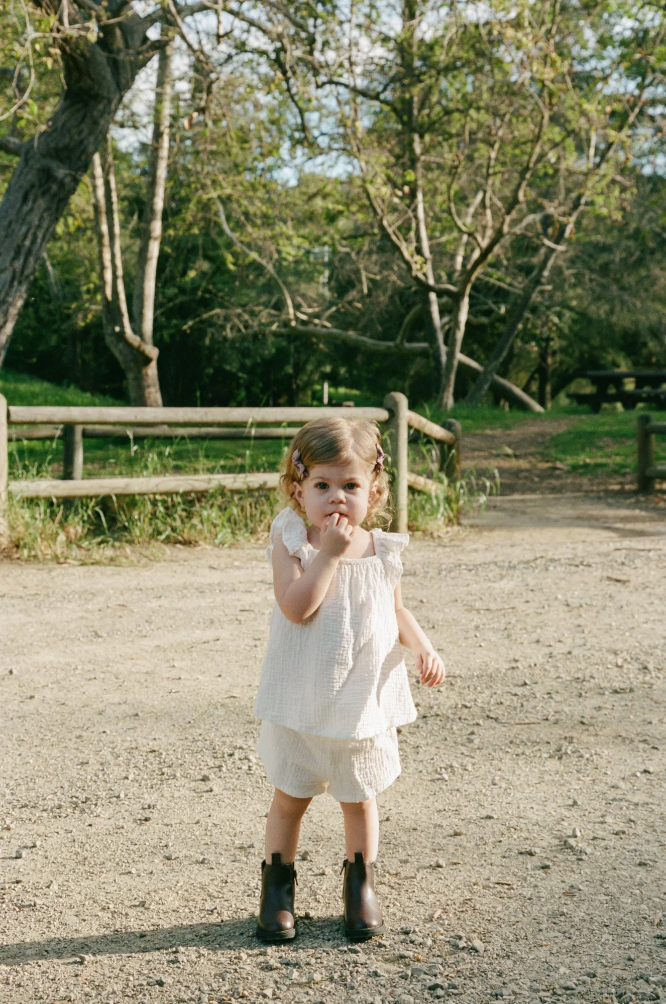 A young girl standing outdoors on a gravel path, wearing a white dress, black boots, and with hair clips in her curly hair, in a park setting with trees and a wooden fence in the background.