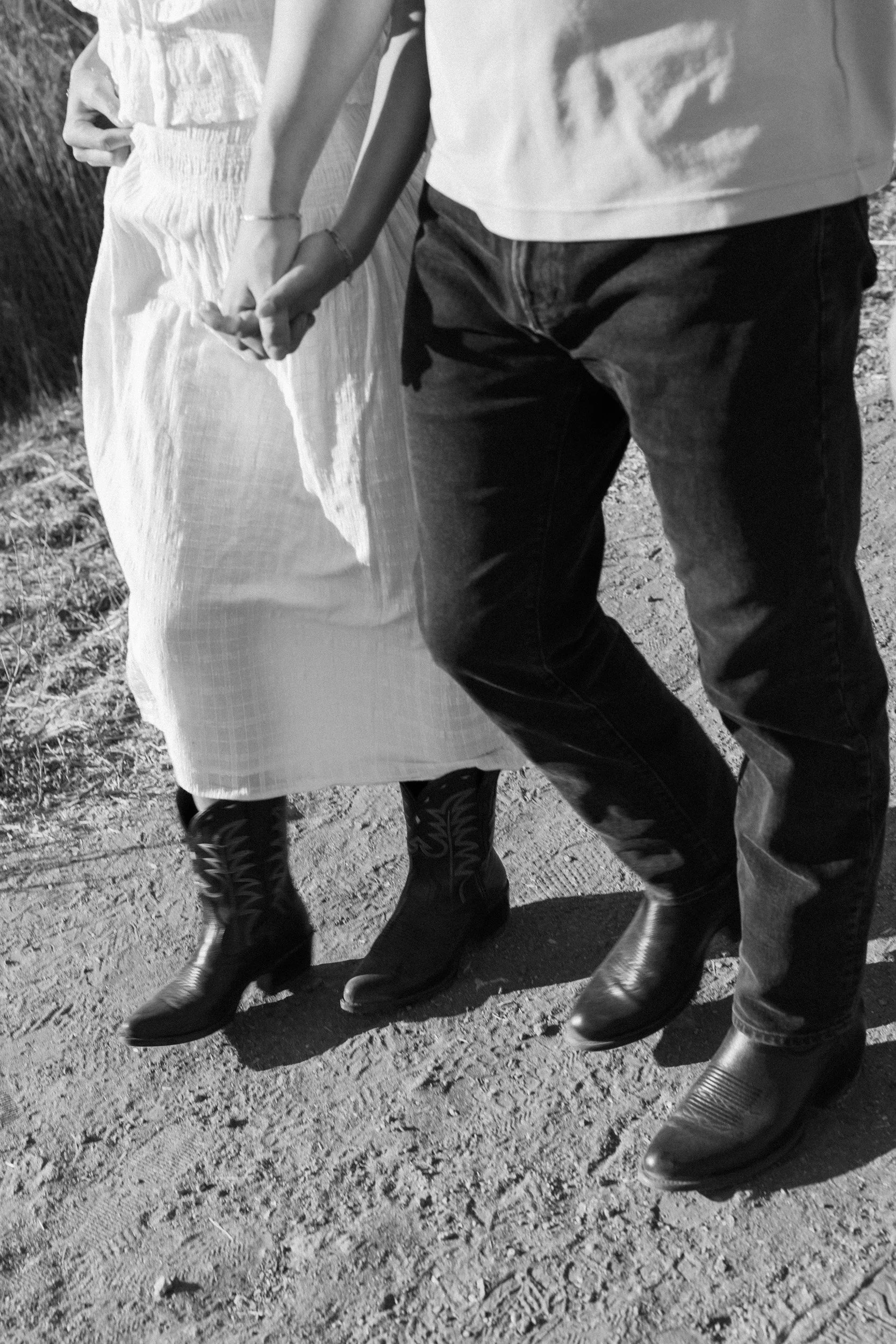 Close-up black-and-white photo of three people standing outdoors on a gravel surface, holding hands, all wearing boots and casual clothing.