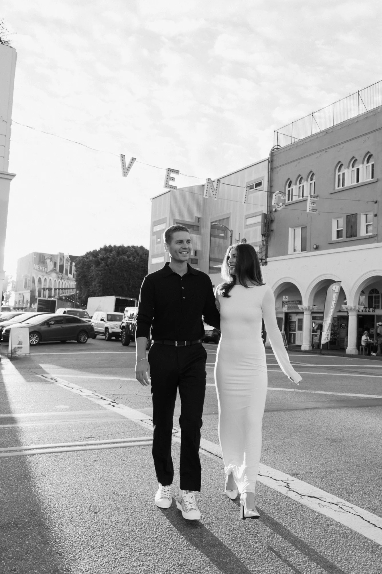 A smiling man and woman walking arm-in-arm on a city street during the daytime. The man is wearing a black shirt and pants with white sneakers, while the woman is in a fitted white dress with high heels. There are buildings, parked cars, and a sign t