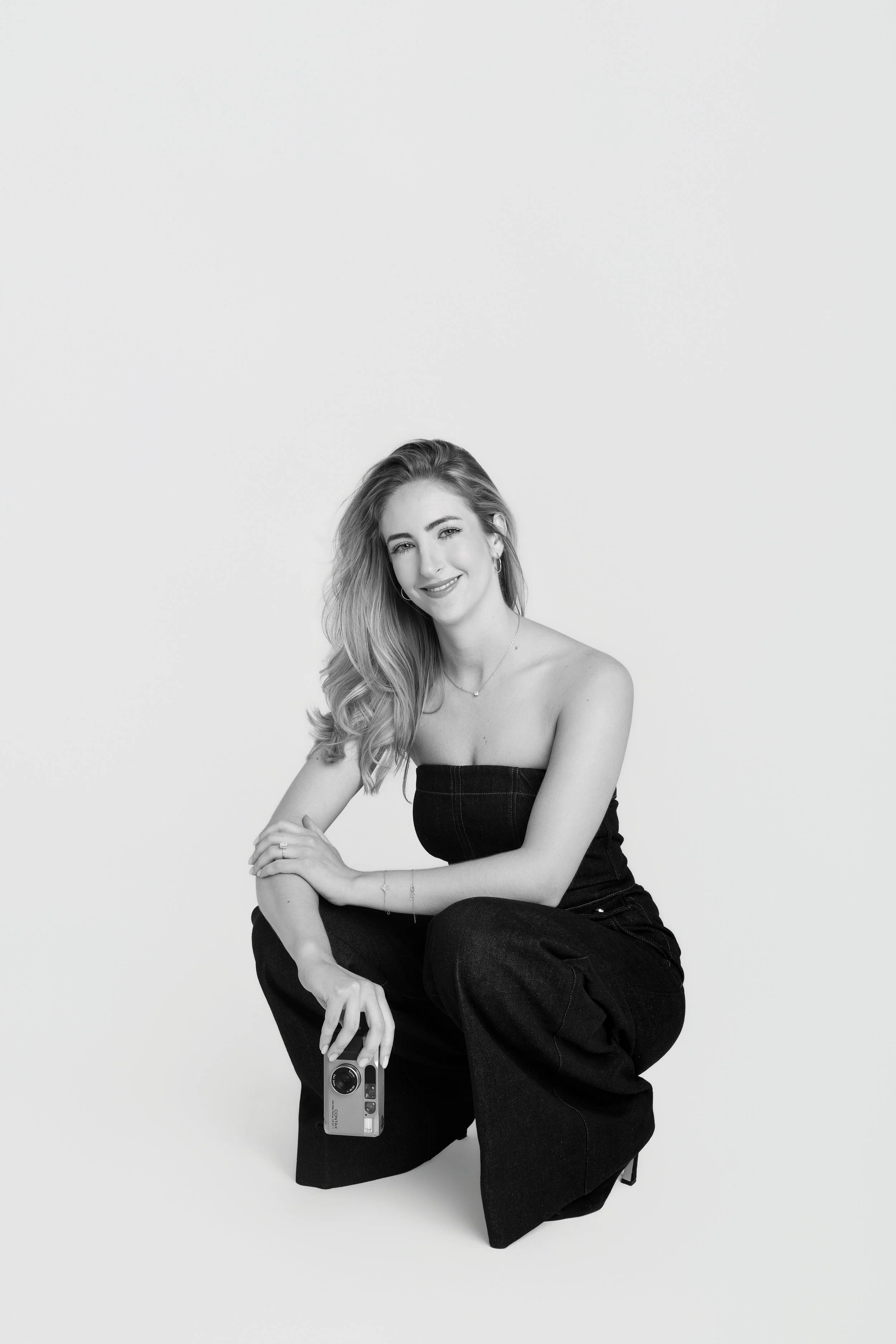 A young woman with long wavy hair, smiling, wearing a strapless top and dark jeans, sitting on the floor against a plain background, holding an old-fashioned camera, with her knees bent and one arm resting on her knee.