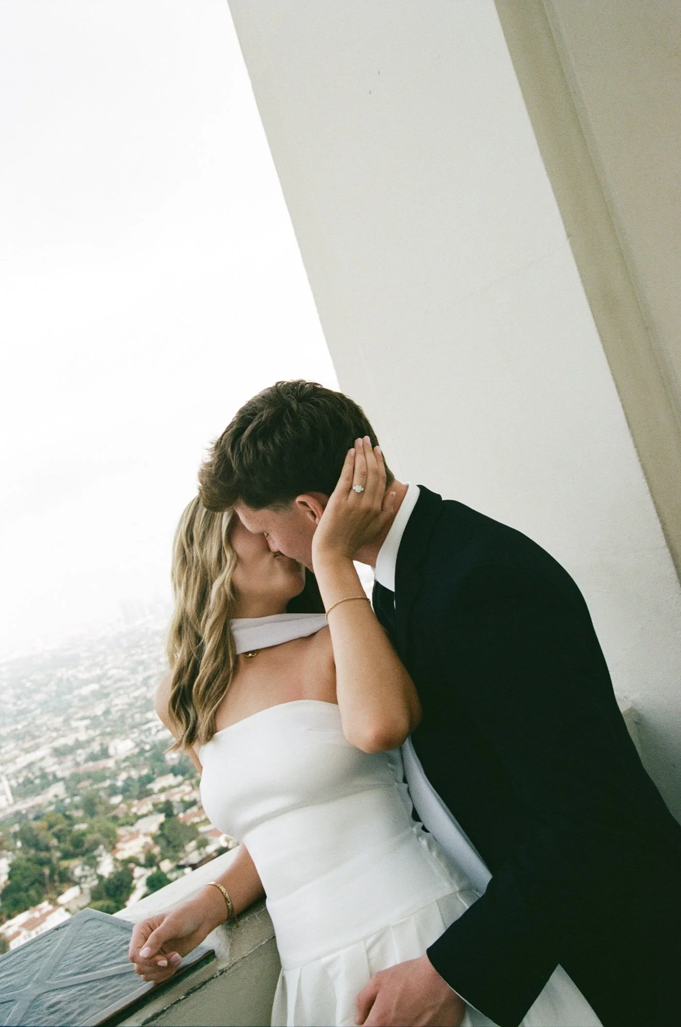 Couple sharing a kiss on a rooftop, with cityscape and water view in the background, during sunset.